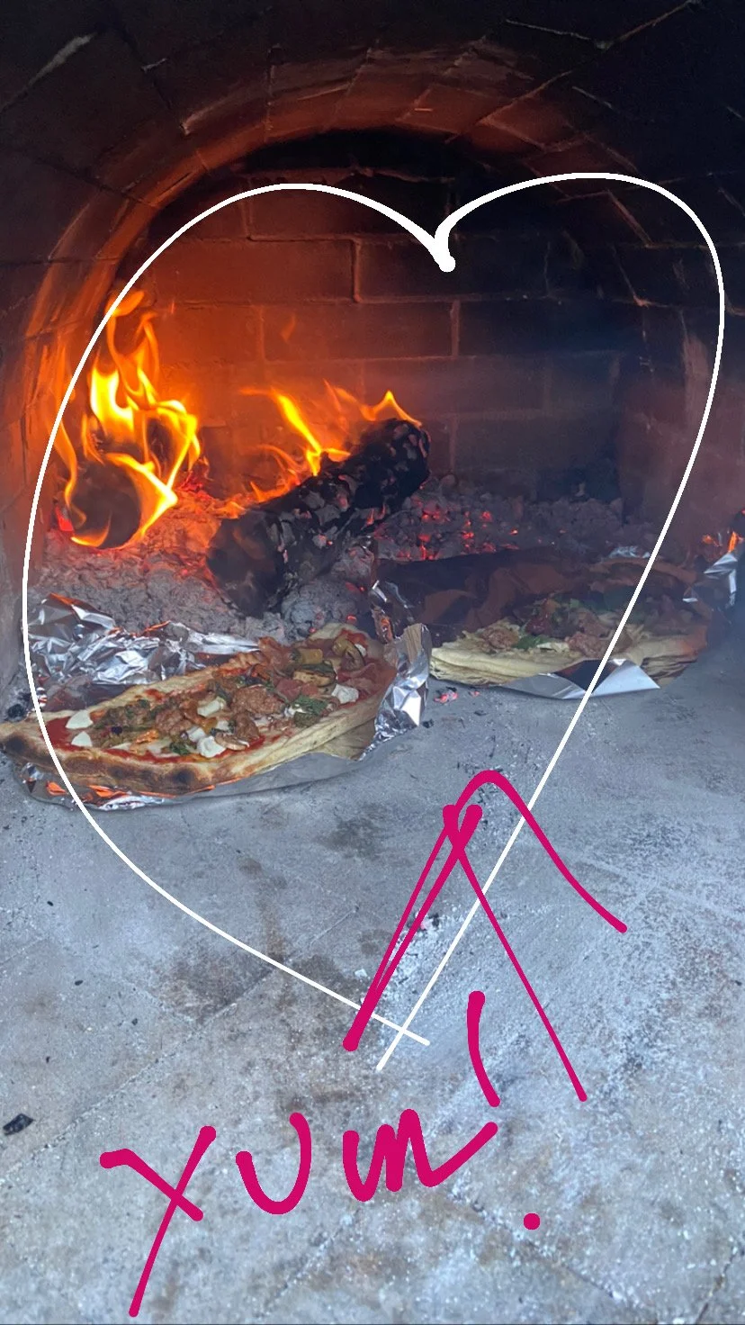 Pizza baking in a wood-fired oven with flames, surrounded by aluminum foil, with a handwritten heart outline and the words 'YUM!' in pink.