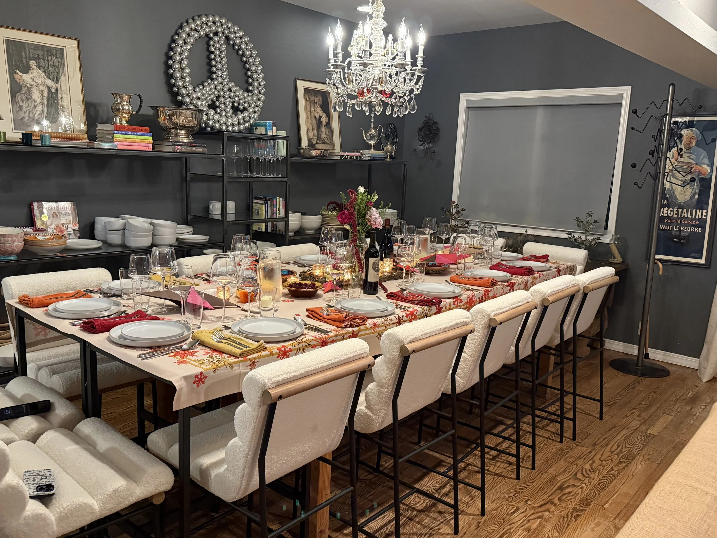 A dining room with a long table set for a meal, featuring white chairs with black frames, a floral tablecloth, wine bottles, glasses, plates, and colorful napkins; a black shelf with bowls, books, and art pieces; a chandelier hangs above, and a peace