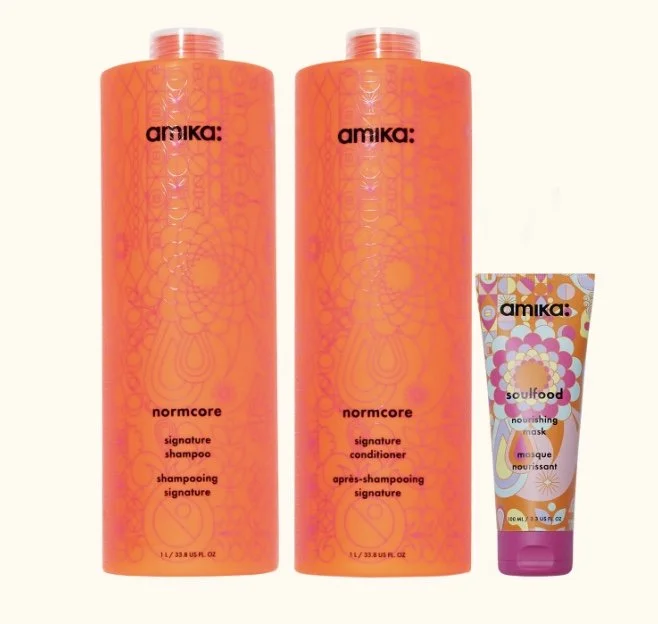 Two large orange bottles of Amika Normcore shampoo and conditioner next to a smaller tube of Amika Soulfood nourishing mask.