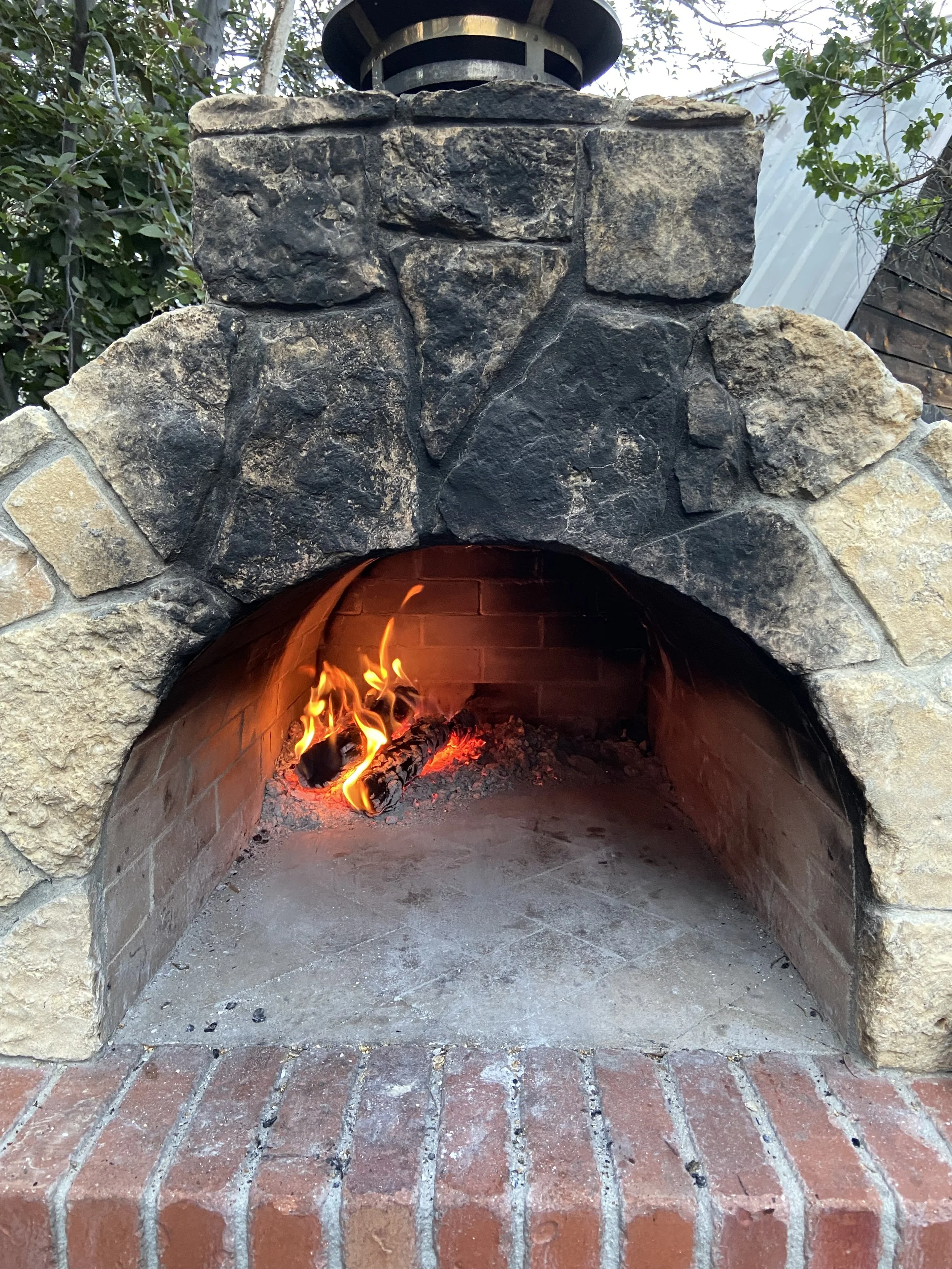 A stone outdoor fireplace with a fire burning inside, surrounded by bricks at the base, and trees and a greenhouse in the background.