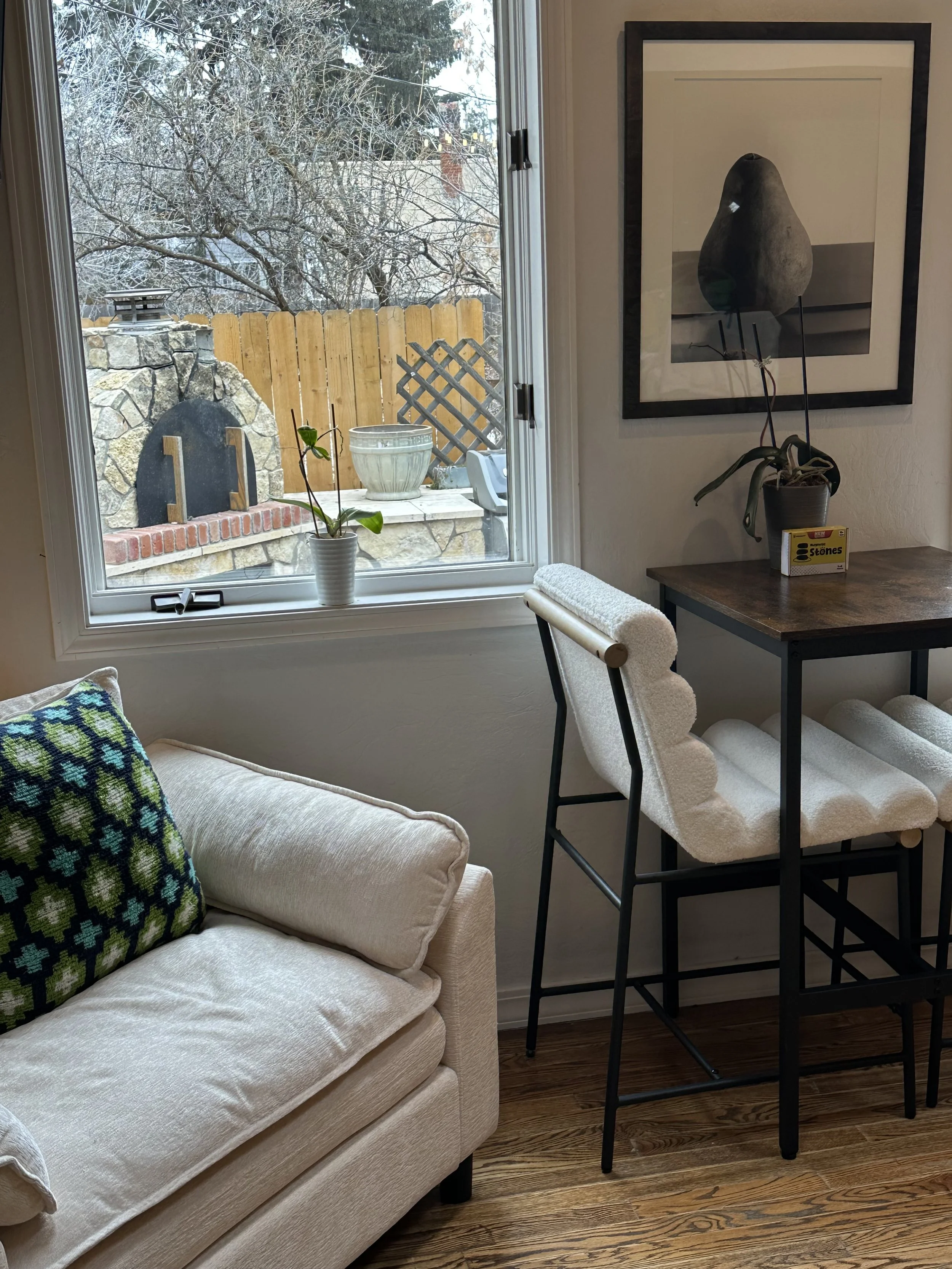 Interior view of a cozy room with a beige sofa with a colorful cushion, a wooden high chair, a small table with a potted orchid, and a wall art piece. Outside the window, there's a snow-covered yard with a stone fireplace and a wooden fence.