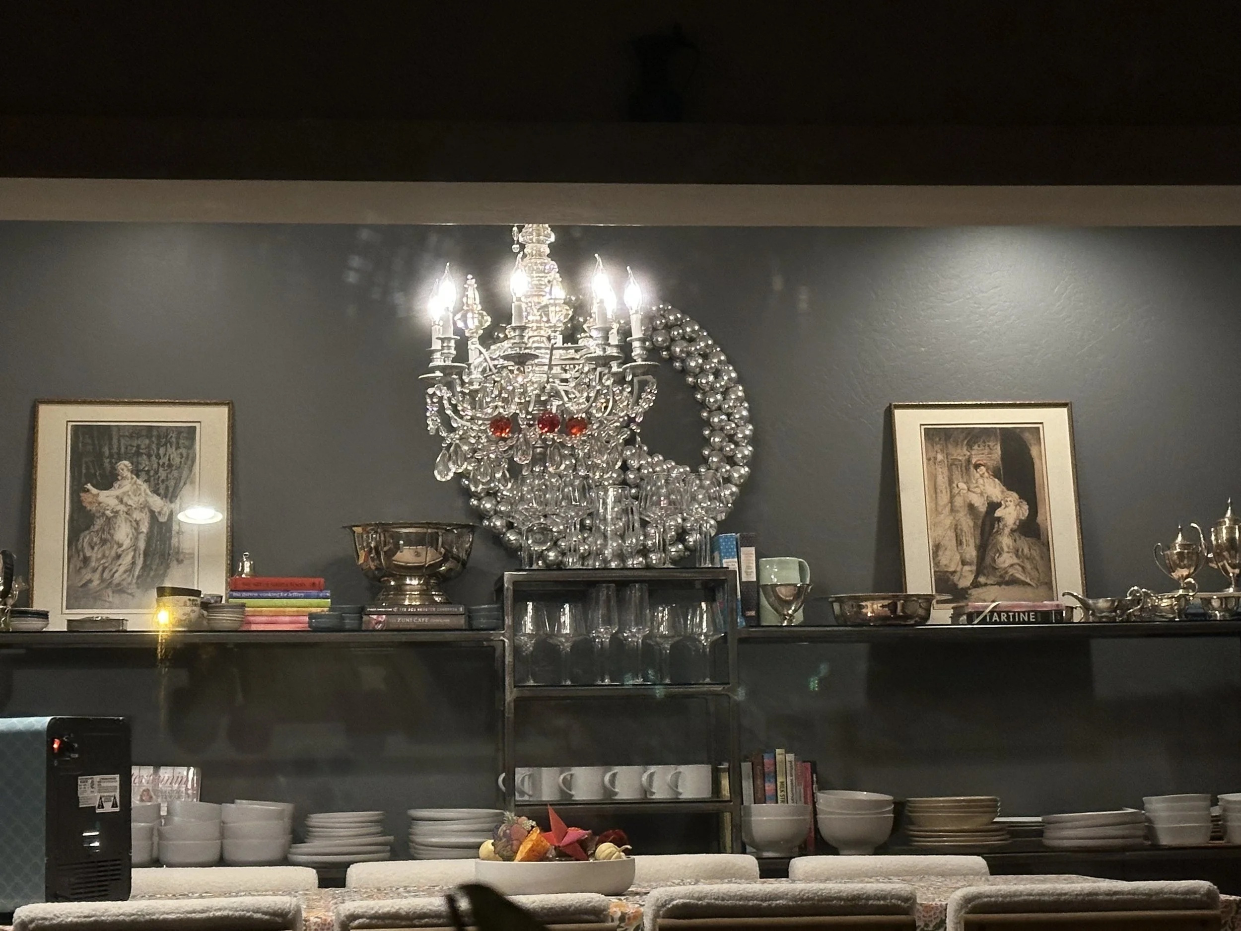 A dark gray wall decorated with framed artwork, a large silver and crystal chandelier, and a shelf filled with bowls, cups, books, and decorative items in the background.