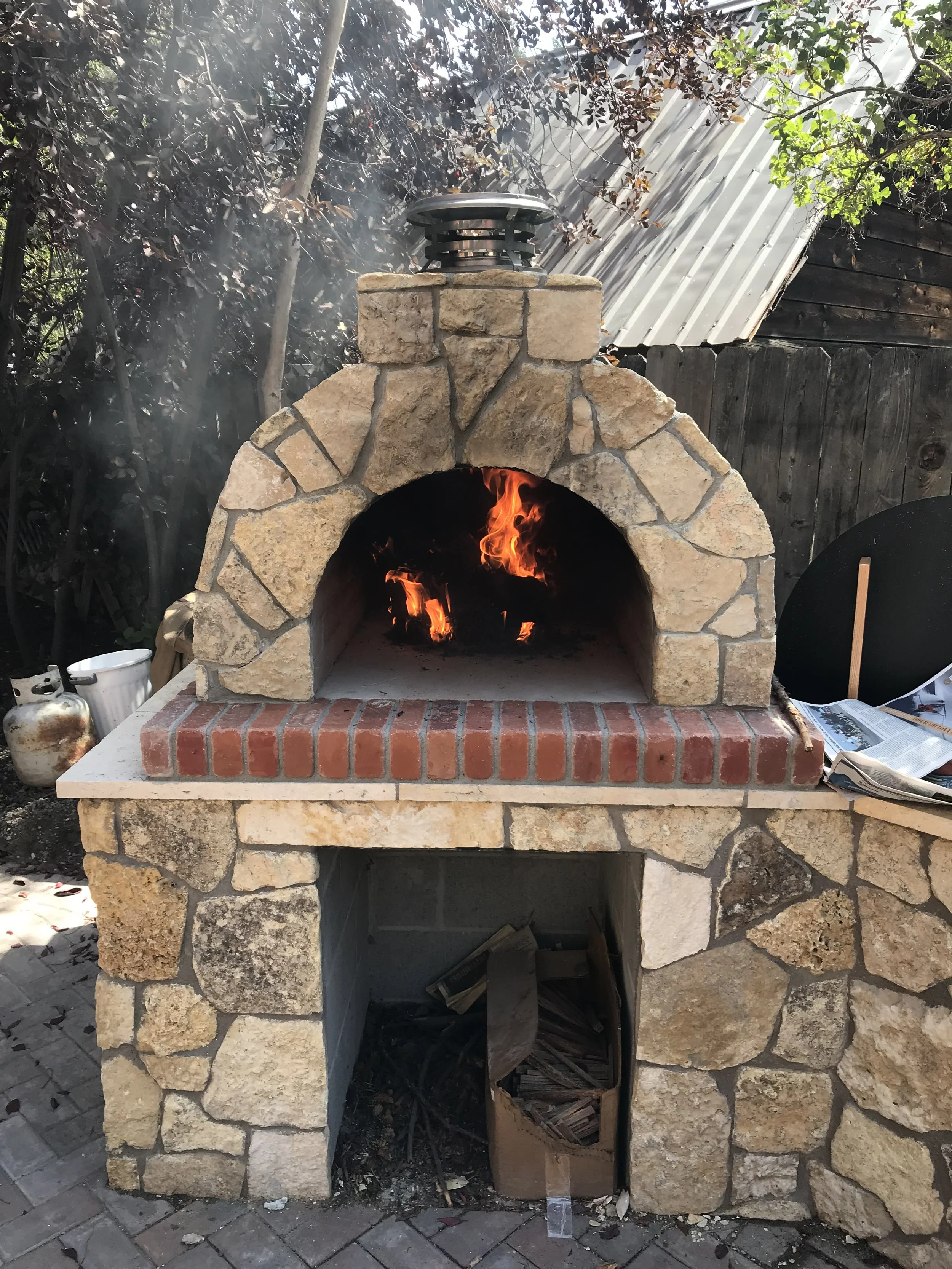 Outdoor stone and brick pizza oven with a fire burning inside, surrounded by trees and rustic buildings.