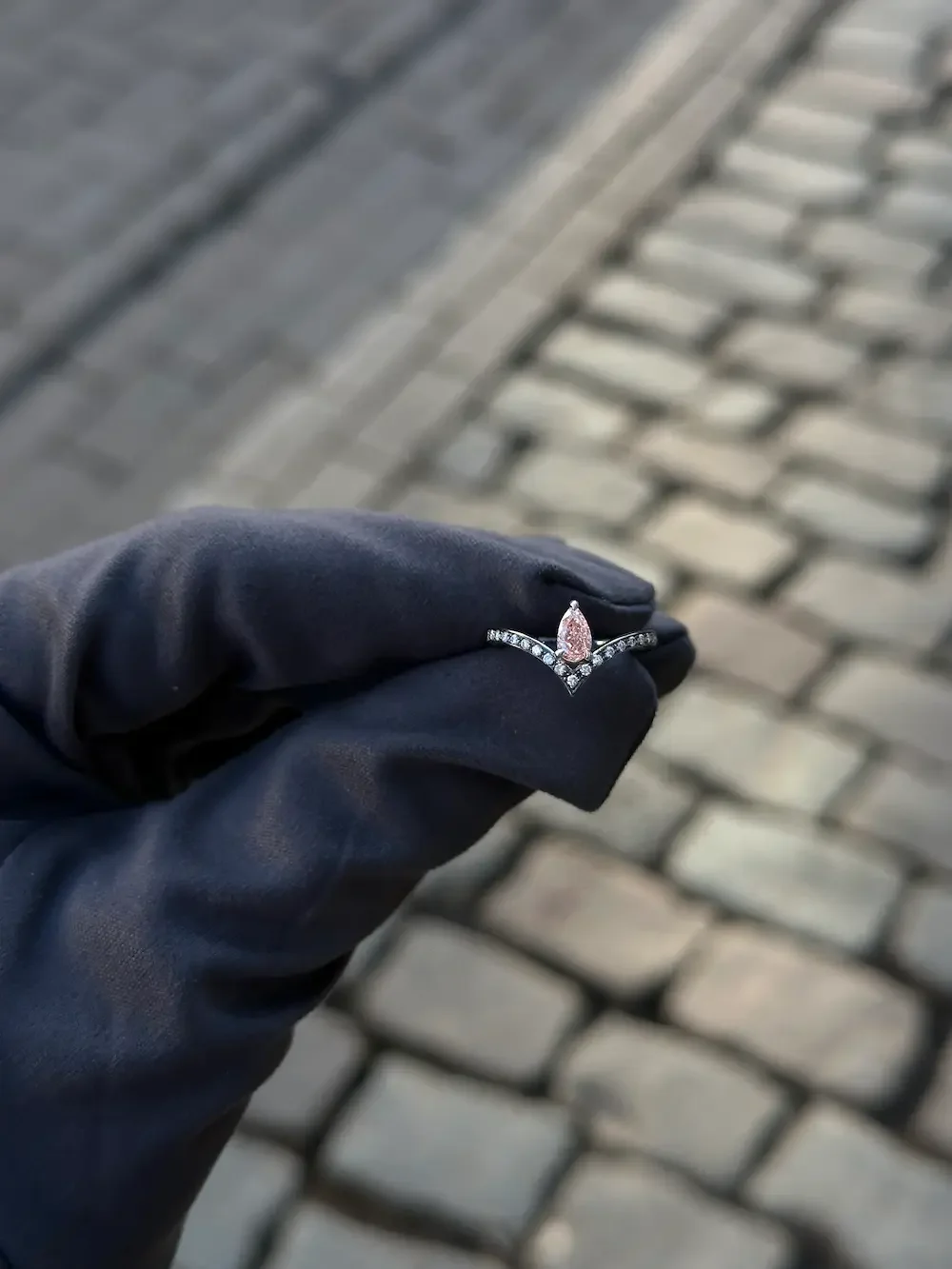 A gloved hand holding a ring with a pink gemstone and small diamonds, against a cobblestone street background.