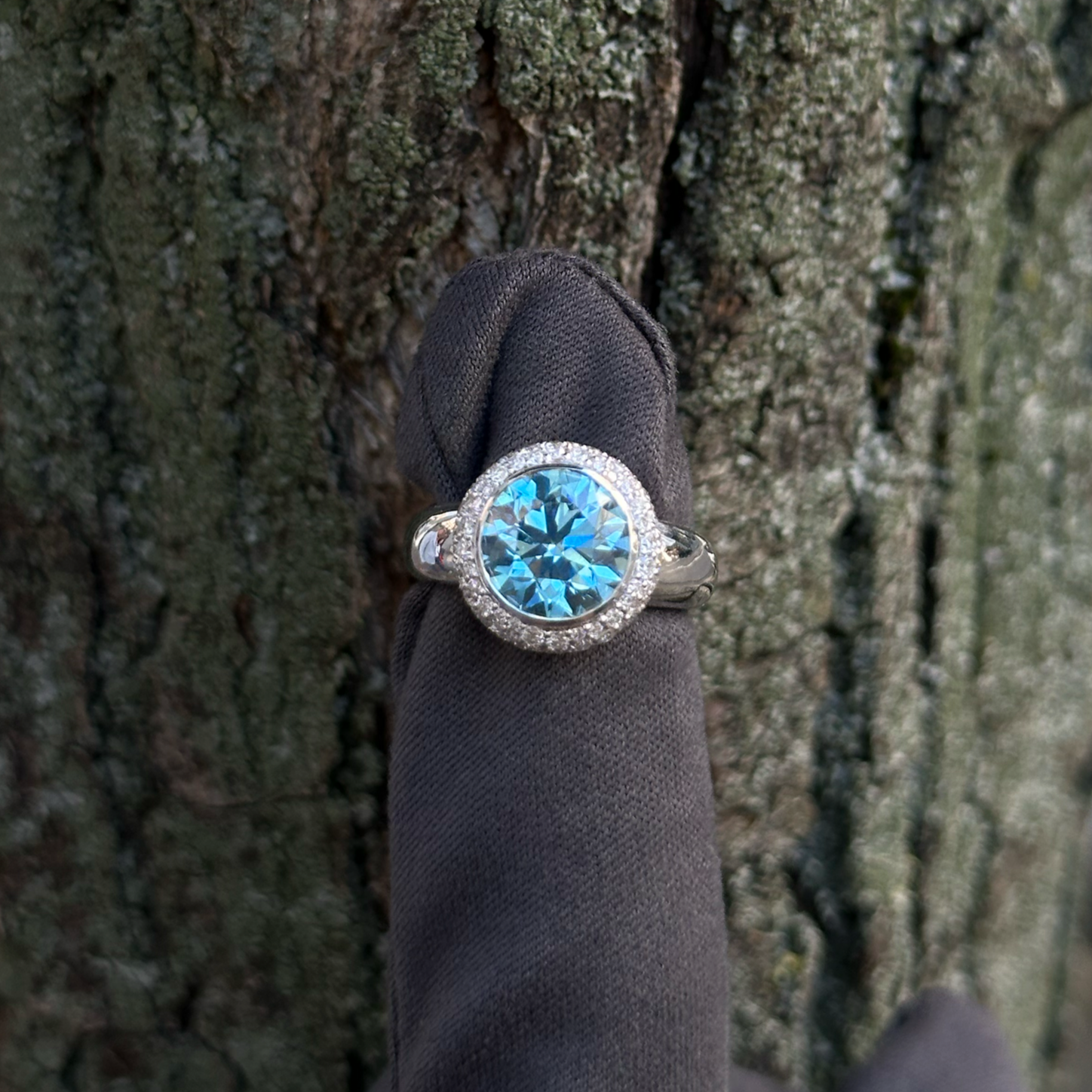Fancy blue vivid diamond ring with a halo of snow set diamonds, worn on a finger covered with a dark grey glove, against the textured bark of a tree.