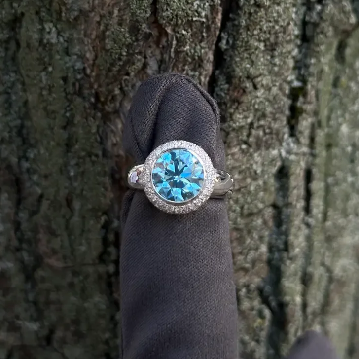 Fancy blue vivid diamond ring with a halo of snow set diamonds, worn on a finger covered with a dark grey glove, against the textured bark of a tree.