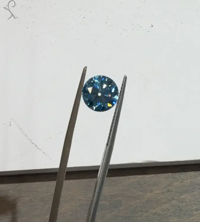 A blue gemstone held by metal tweezers over a flat surface with a white background.