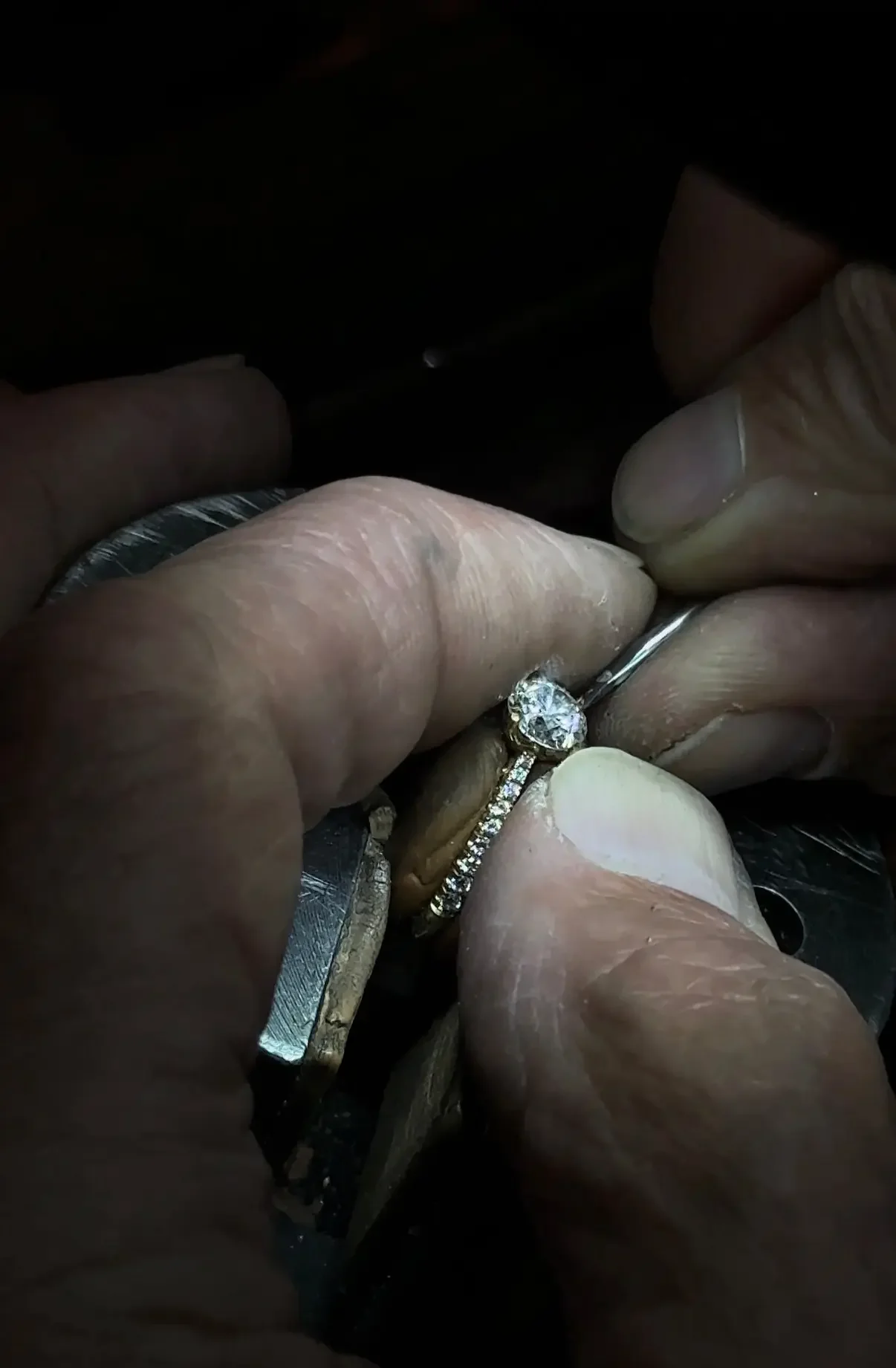 Close-up of hands working on a piece of jewelry, specifically a ring with a large diamond center stone and smaller diamonds on the band.
