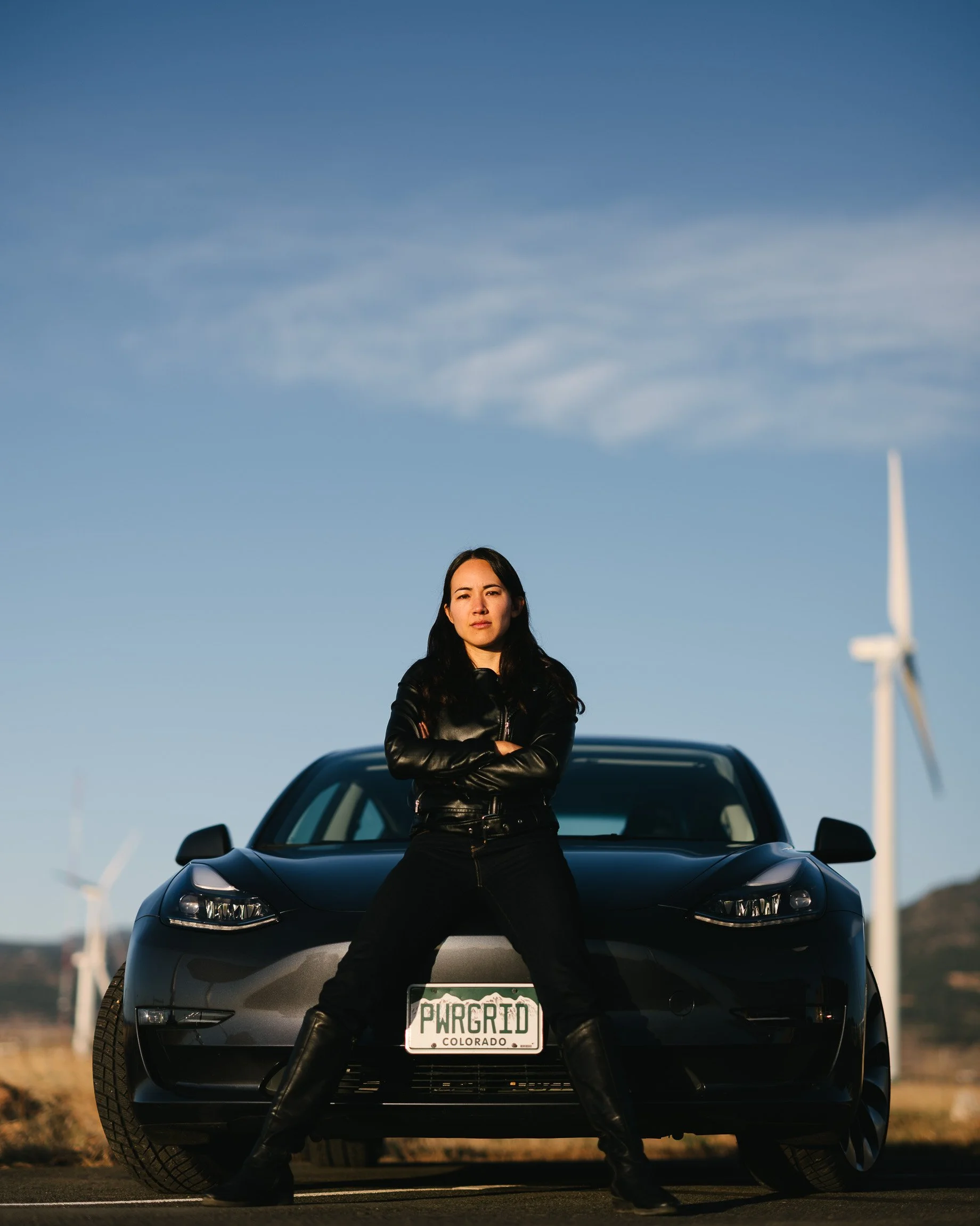 Kyri Baker stands in front of her black Tesla, dressed in black in an aggressive pose, an NREL wind turbine behind her and her PWRGRID license plate.