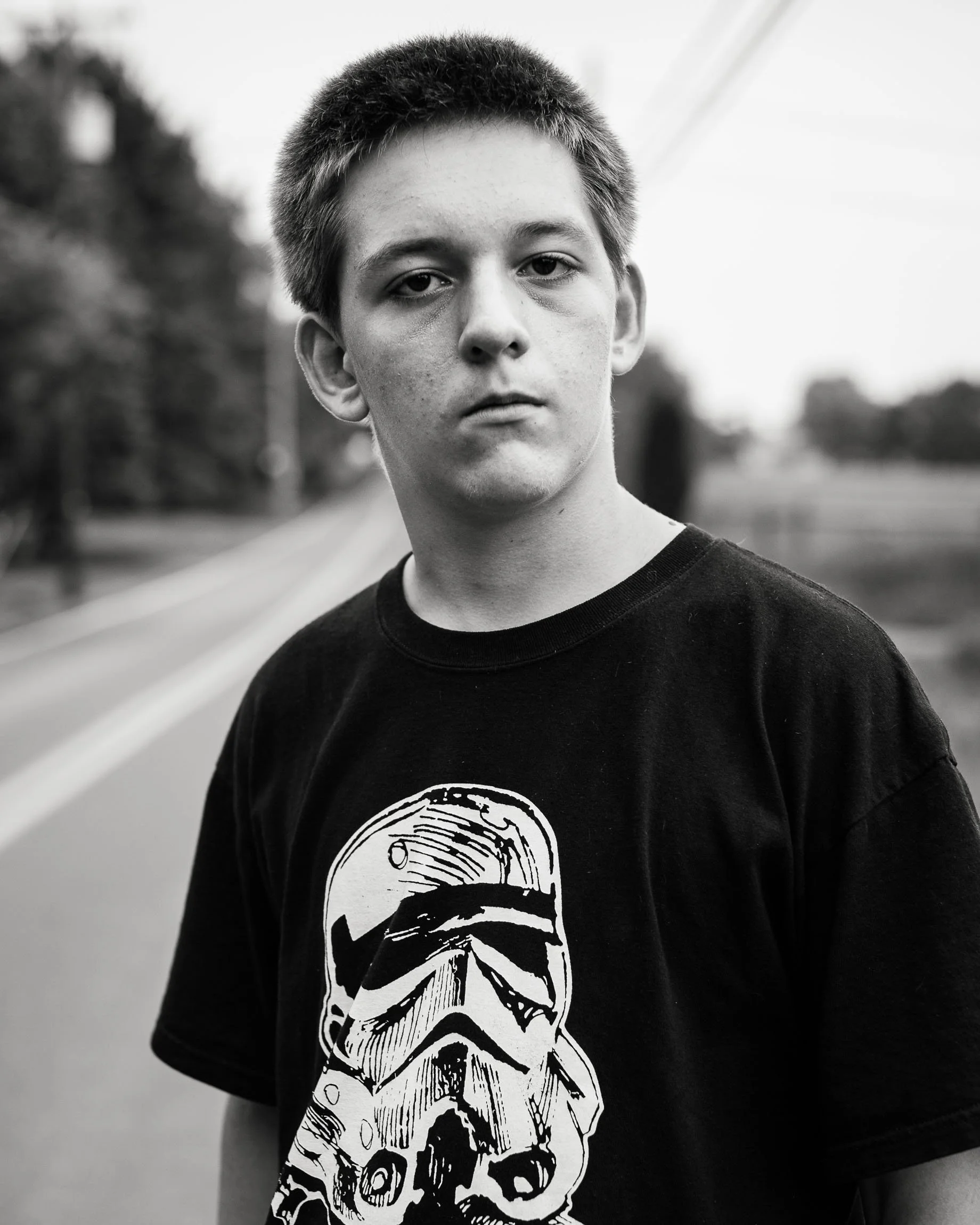 Head and shoulders monochrome portrait of a young man standing on a rural road with a black t-shirt with a Star Wars stormtrooper, his own expression mirroring that of the character.