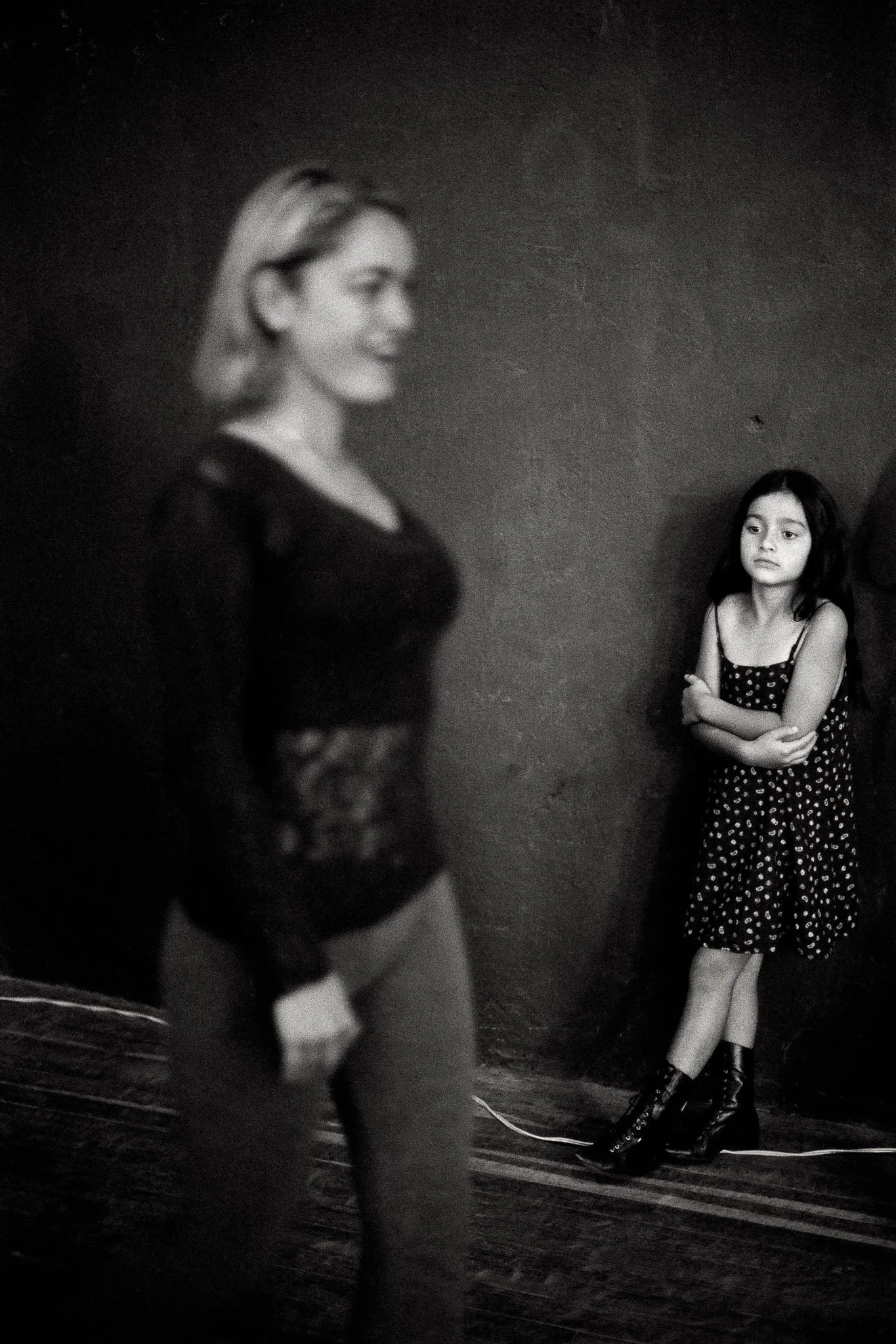 In a dark image of a beauty queen practice session, a young woman practices walking but the focus is on a young girl clutching herself in the background, in focus, in a black patterned dress and black boots. It speaks to the process of acculturation.