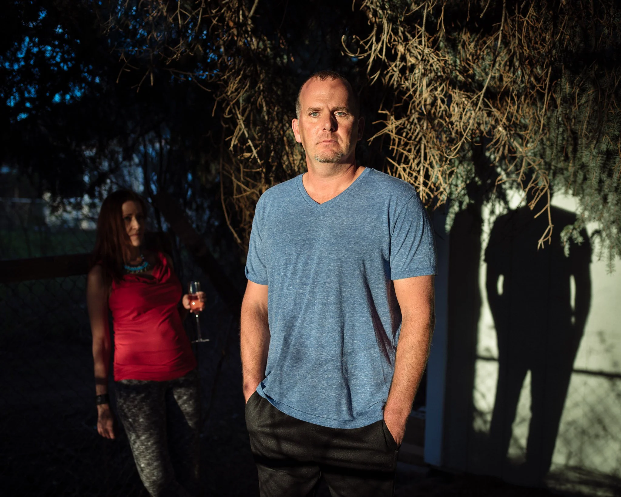 Middle aged man with receeding hairline and chin stubble standing in a patch of sunlight in his dark yard, a woman in the shadows behind him holding a glass of wine. He looks directly at viewer.