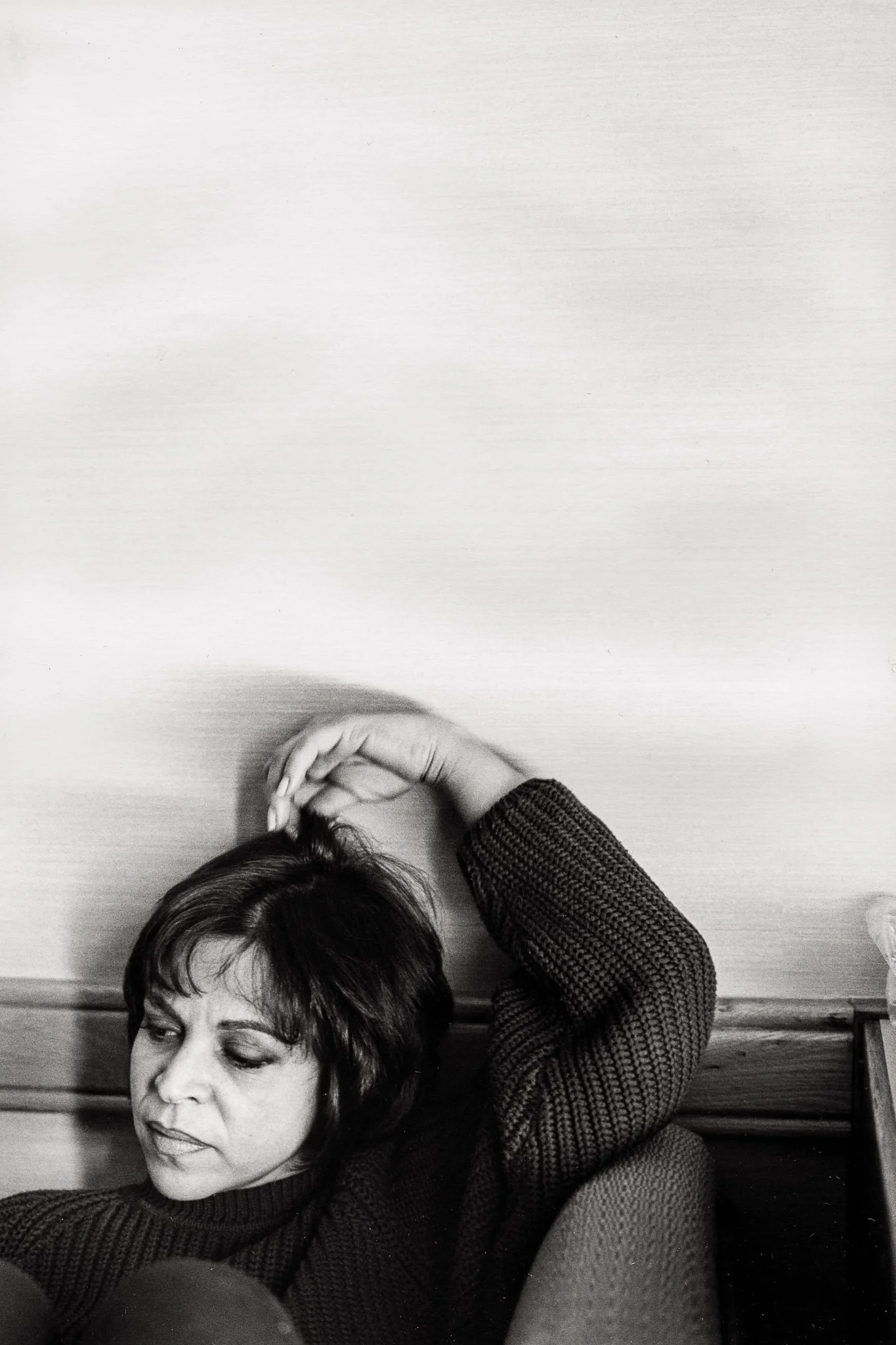 A woman sits on the lower corner of the frame, tightly cropped, knees visible, her arm upraised and bent over her head, with a somewhat defeated expression. Frame is mostly negative space, she is small and at the bottom.