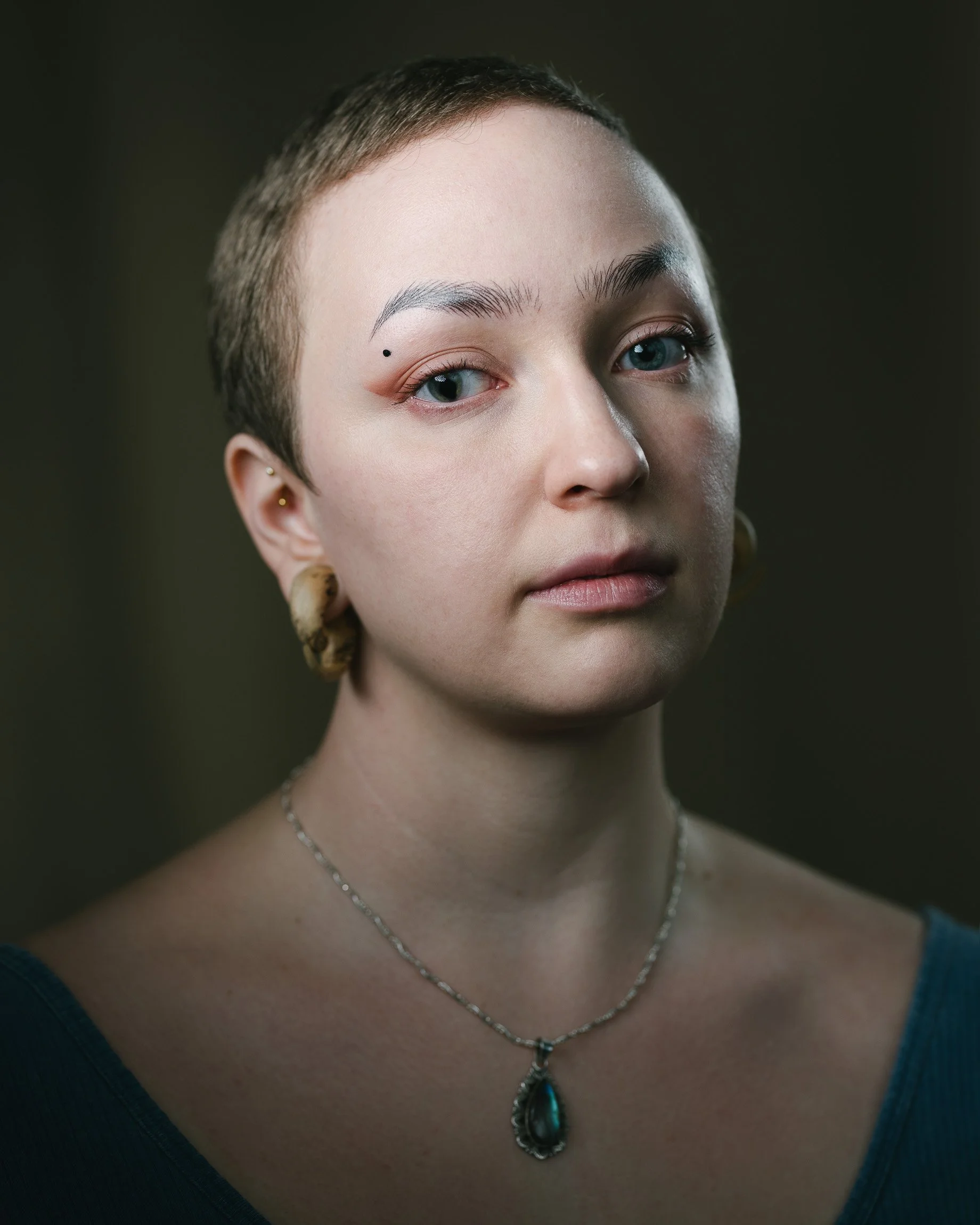 Portrait of a young woman with extremely short hair, decal eyebrows, a birthmark above her eye which has a non-circular iris, her head tilted back, dramatic lighting, a neckless with a gemstone around her neck.