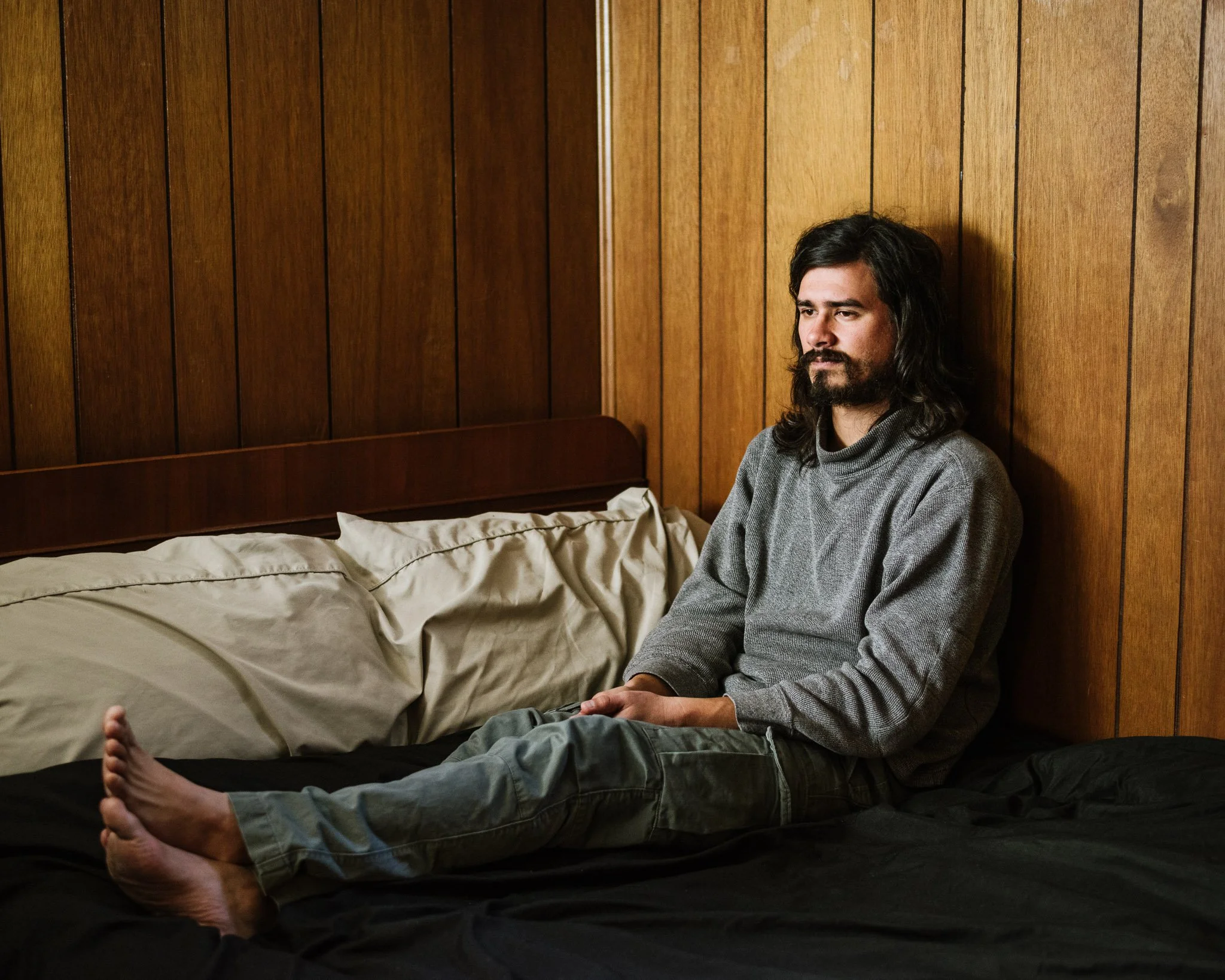 A young white man with long black hair and a full beard sits on the bed of his basement bedroom leaning against the 70s wood paneling in a loose gray sweater, cargo pants and bare feet.