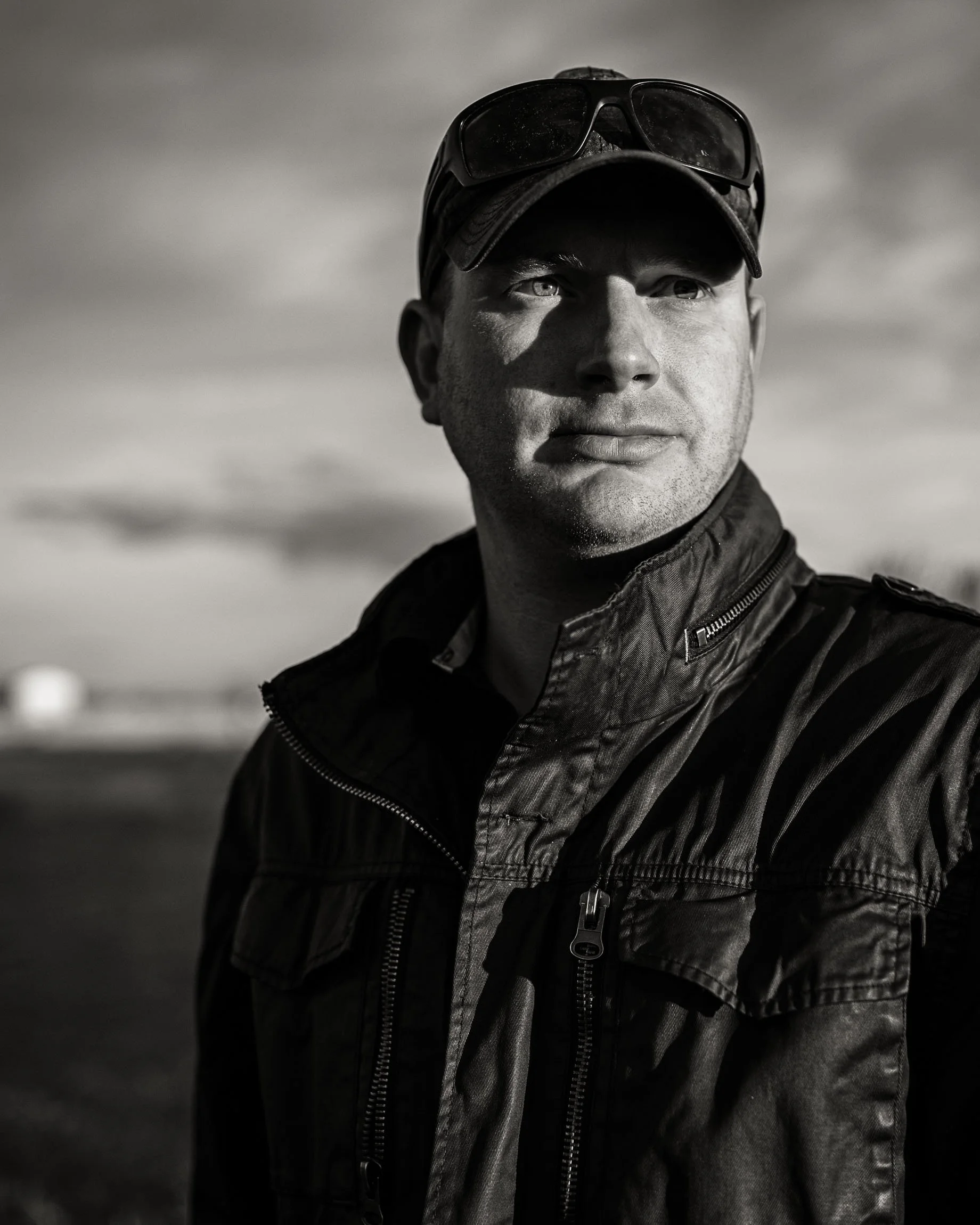 Monochrome image of a man on the prairie in a leather jacket, baseball hat with wrap around sunglasses on his head, looking into distance, face in hard mixed shadow.