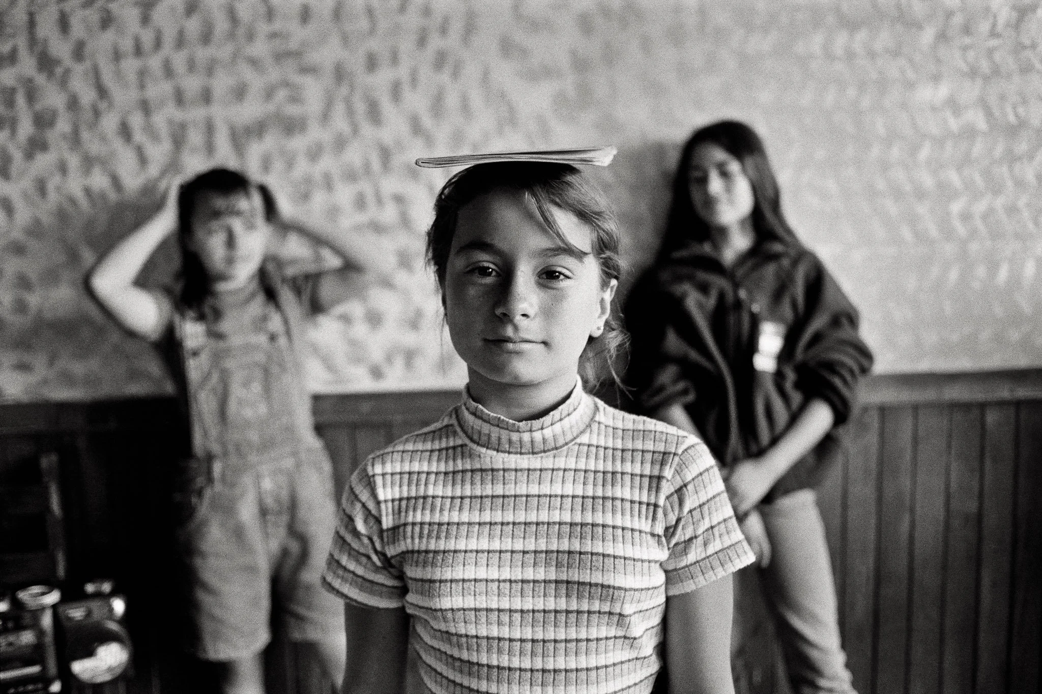 A girl stands with a small book on her head, practicing her posture, centered in the frame, looking at the camera with a small smile. Two other girls are out of focus n the background with a boombox. The feel is innocent and aspirational.