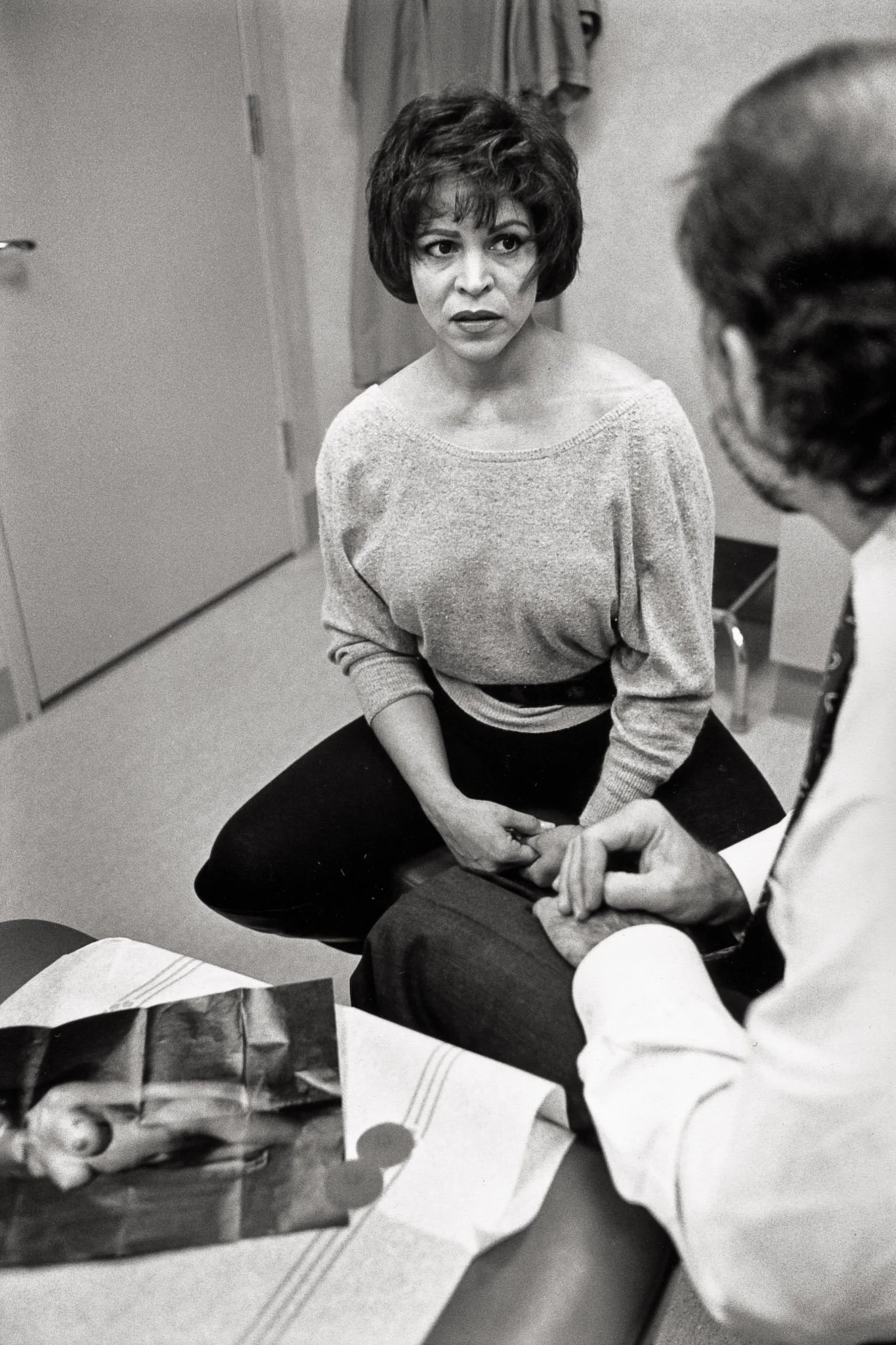 A woman consults with her plastic surgeon after a bilateral mastectomy. In the foreground is a picture of a woman with firm full breasts ripped from a magazine, along with two prosthetic nipples. He explains the limits to what he can do.