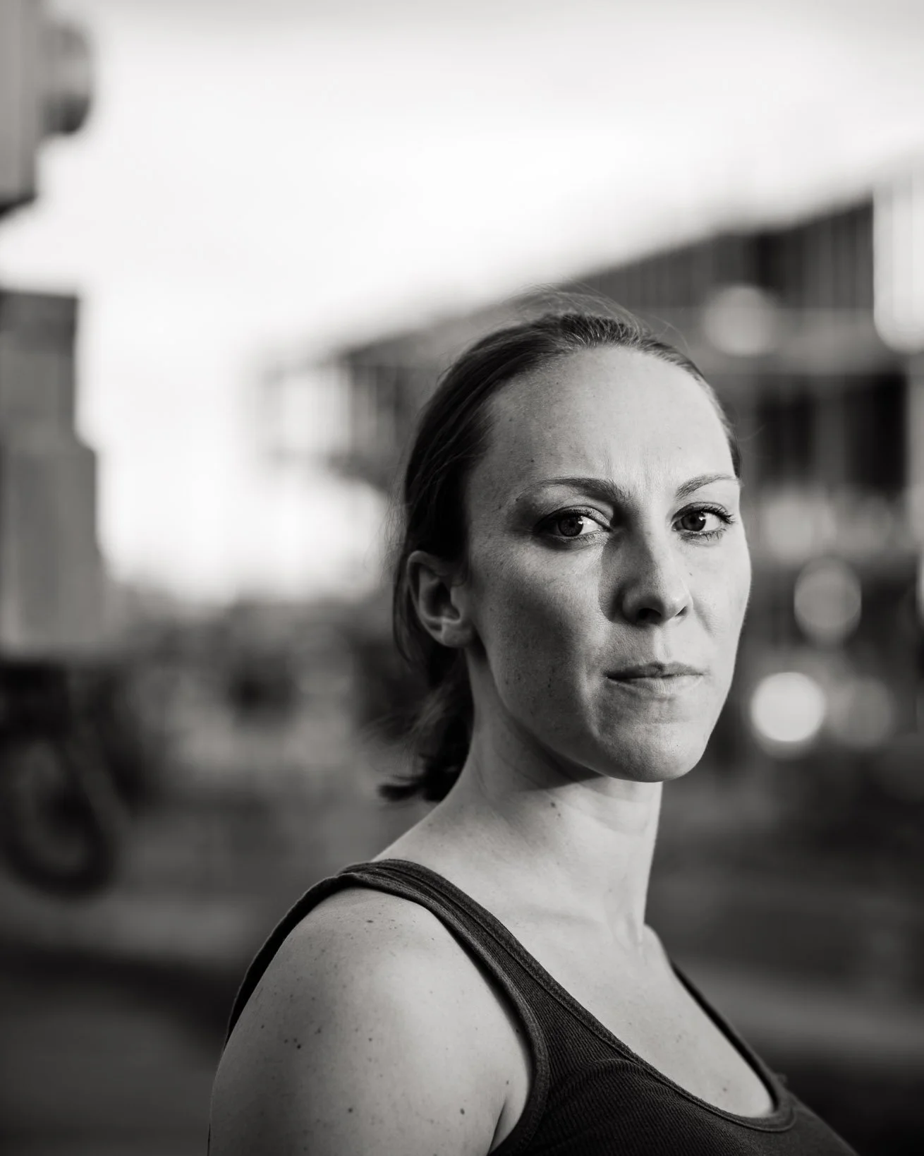 Dramatic monochromatic portrait of a woman in a tank top, closeup head and shoulders, in an urban area in early evening. She has a watchful, knowing gaze.