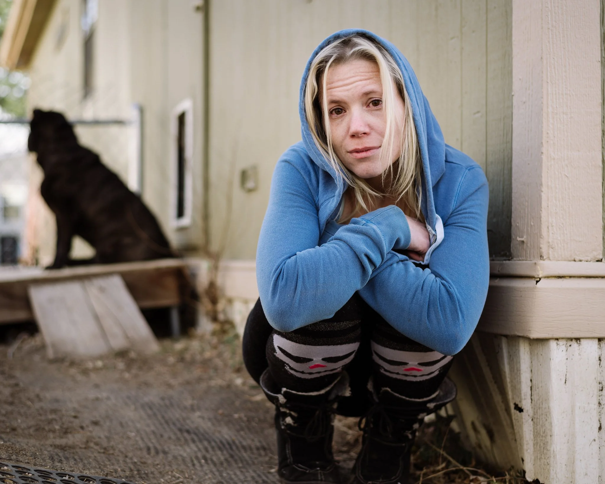A young blond woman in a blue hoodie, stockings with skulls, and boots squats next to her house in a state of disrepair. Her dog is in the background.