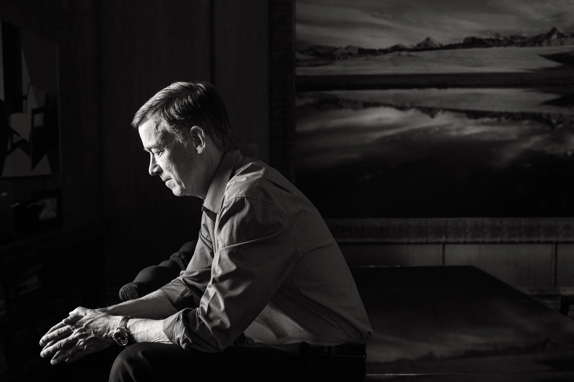 John Hickenlooper sits in the darkness of his office with his eyes closed, elbows on his legs, his hands together before him.