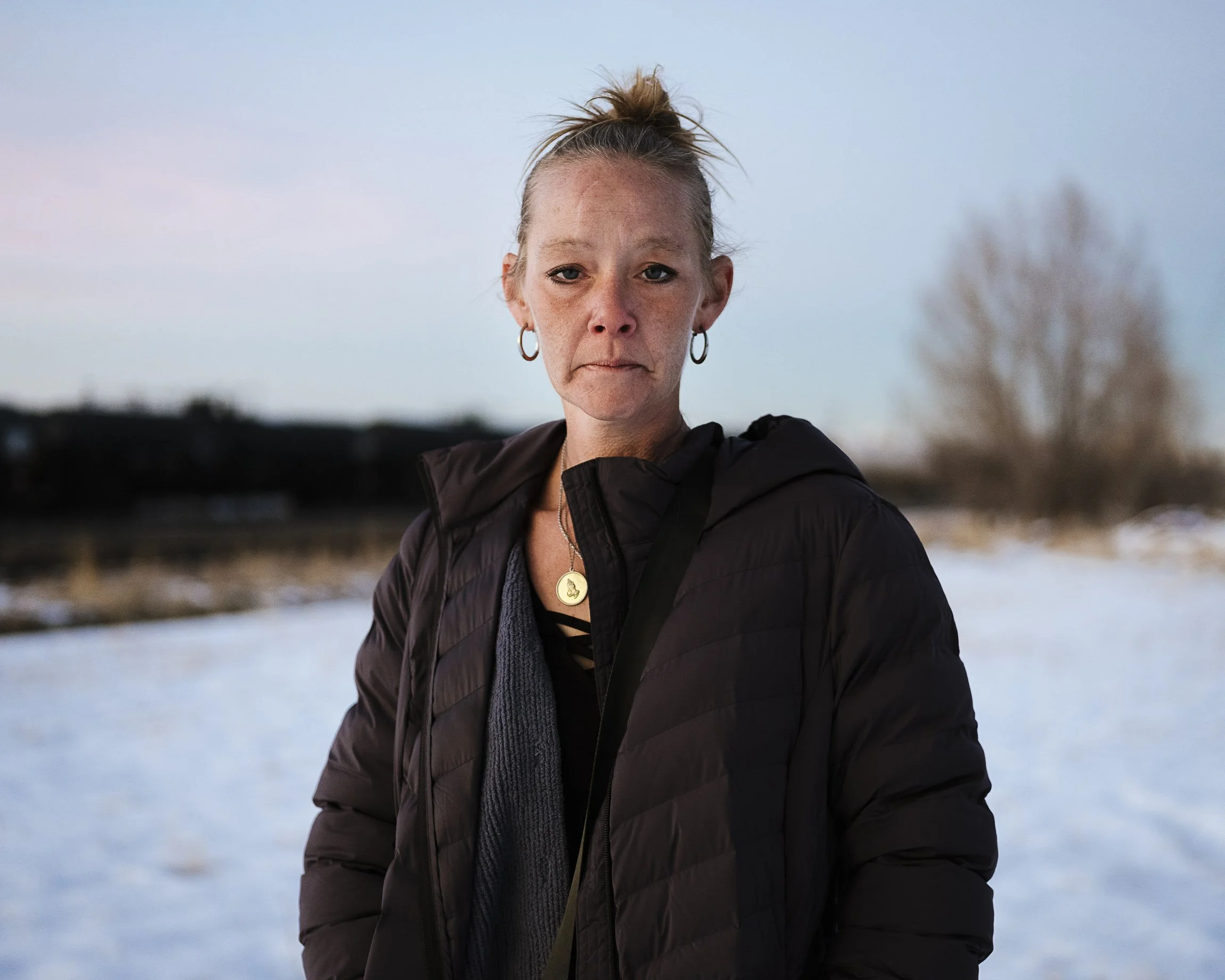 A middle-aged white homeless woman in her coat at dusk in winter in the middle of the frame. Her hair is pulled back, she looks, tired, resigned. A gold St. Christopher's pedant around her neck.