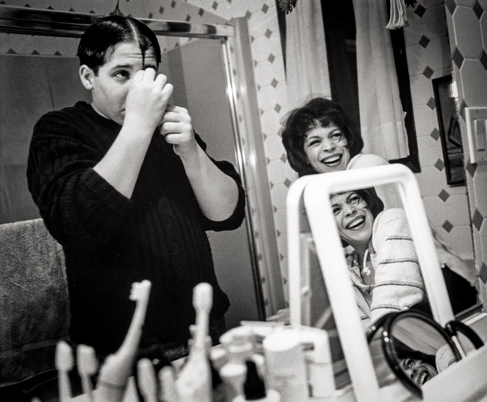 A teenage boy plays with his hair in a silly fashion while his mother watches, reflected in multiple mirrors.