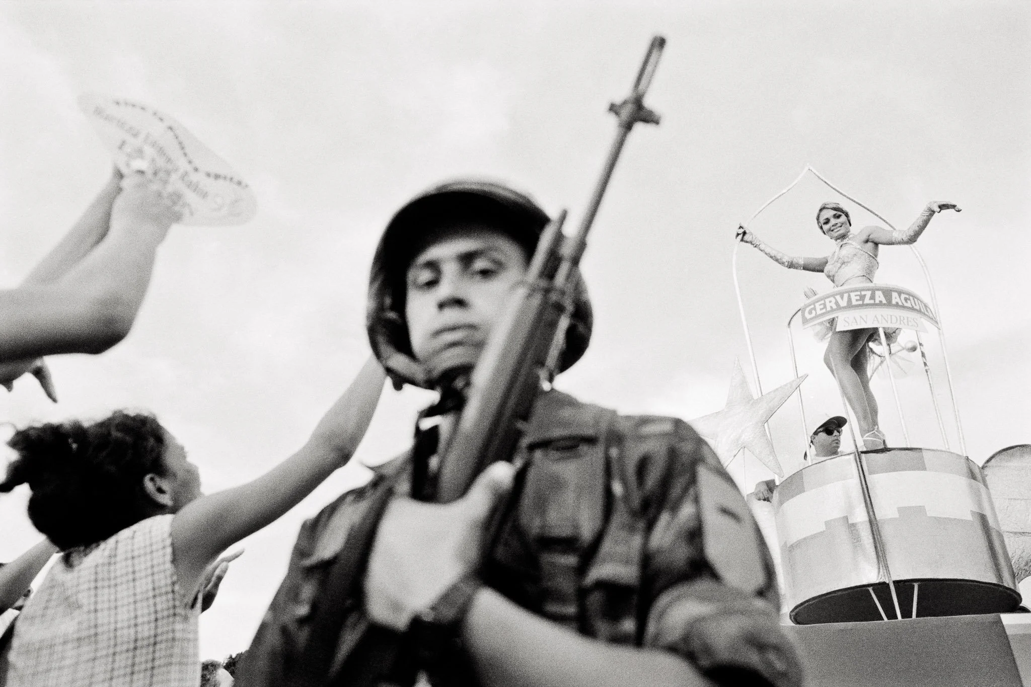 A beauty queen dances on a parade float, small in the background. In the foreground looms a soldier with a rifle, looking at viewer, with suggestions of adoring onlookers to the side. She is the focus, but is visually almost an afterthought, tiny.