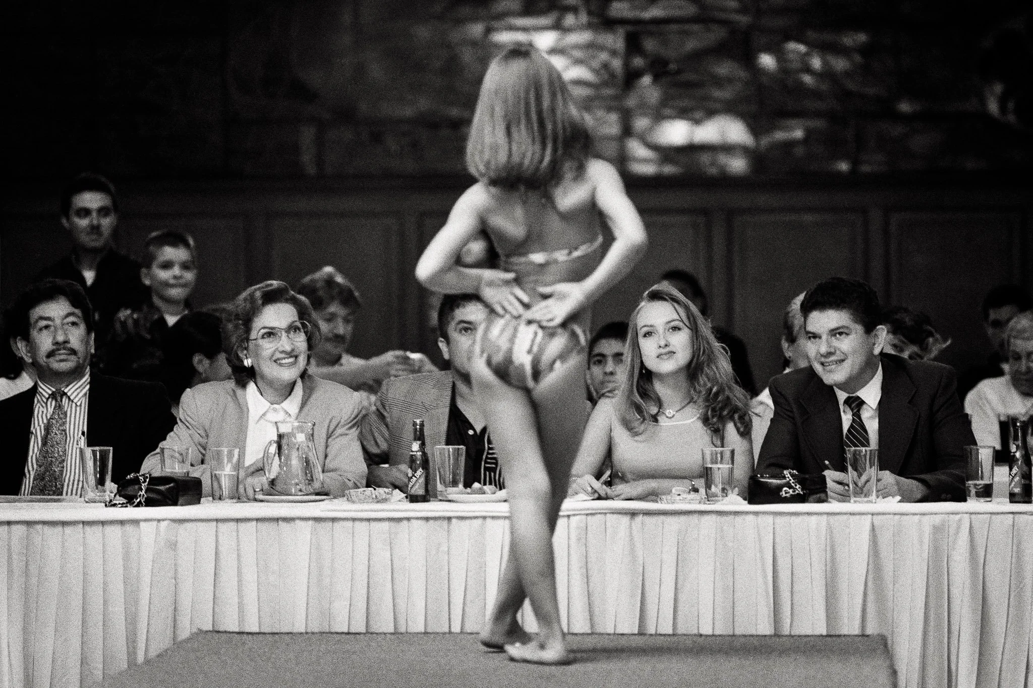 A four year old beauty contestant stands before judges in a pageant in her bikini, feet crossed, arms in the small of her back. Viewed from behind, focused on the judges' expressions.