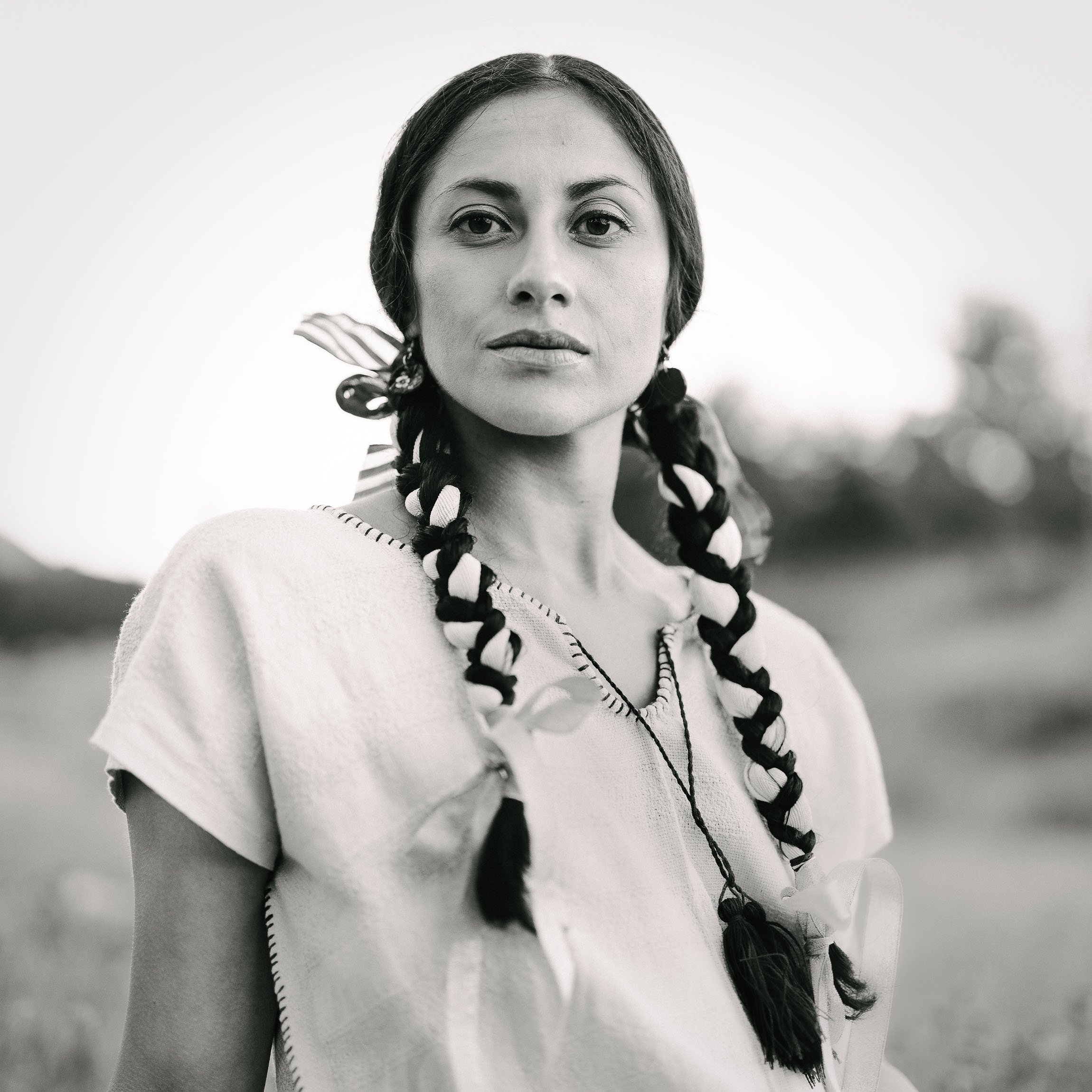 Monochrome square image of young Mexican woman in a meadow in subtle indigenous dress looking at viewer with strength and dignity.