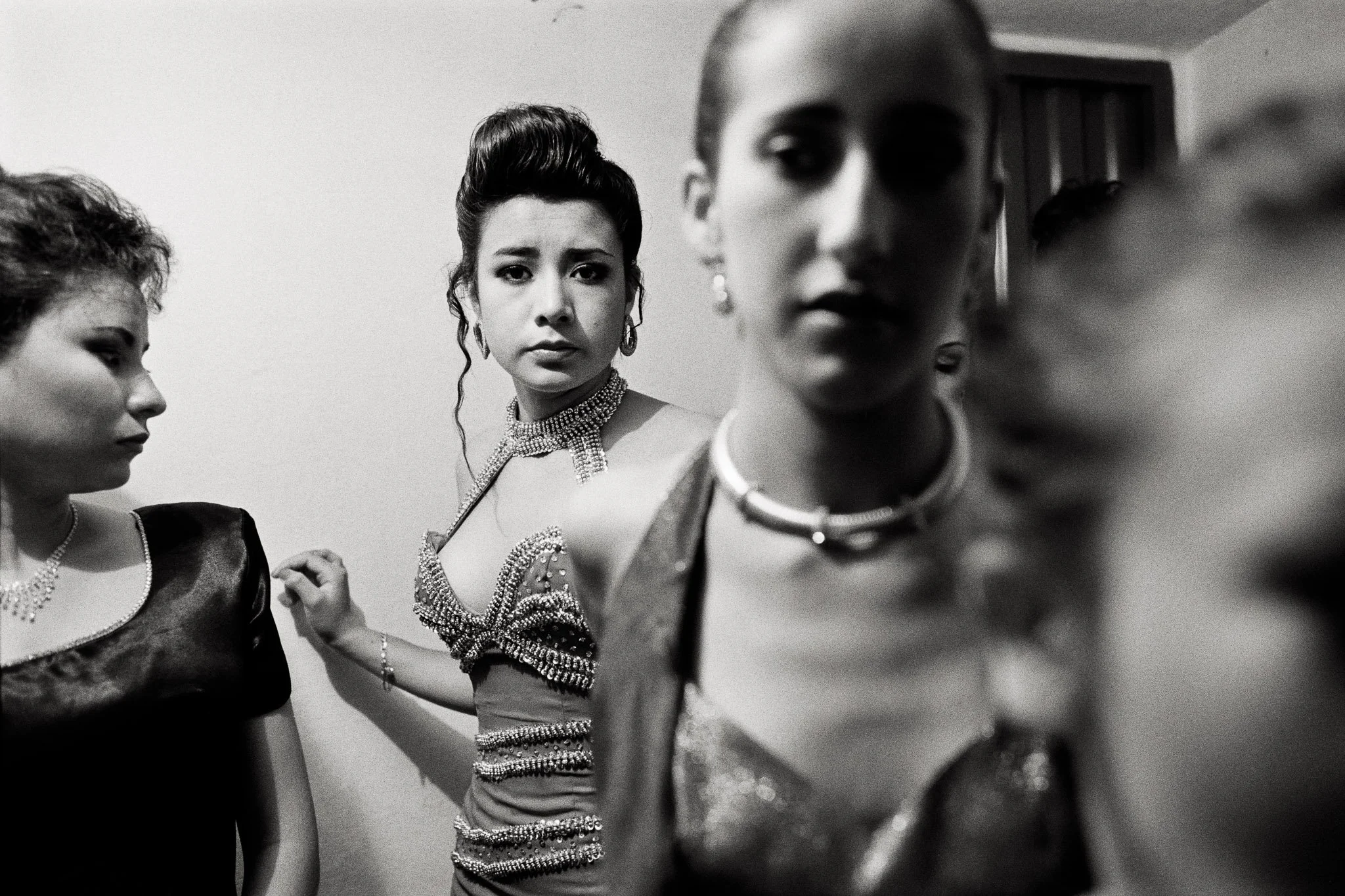 Backstage at a Colombian beauty pageant, women crowded in evening wear. One woman looks directly at the viewer, while another looks at the first with a critical gaze.