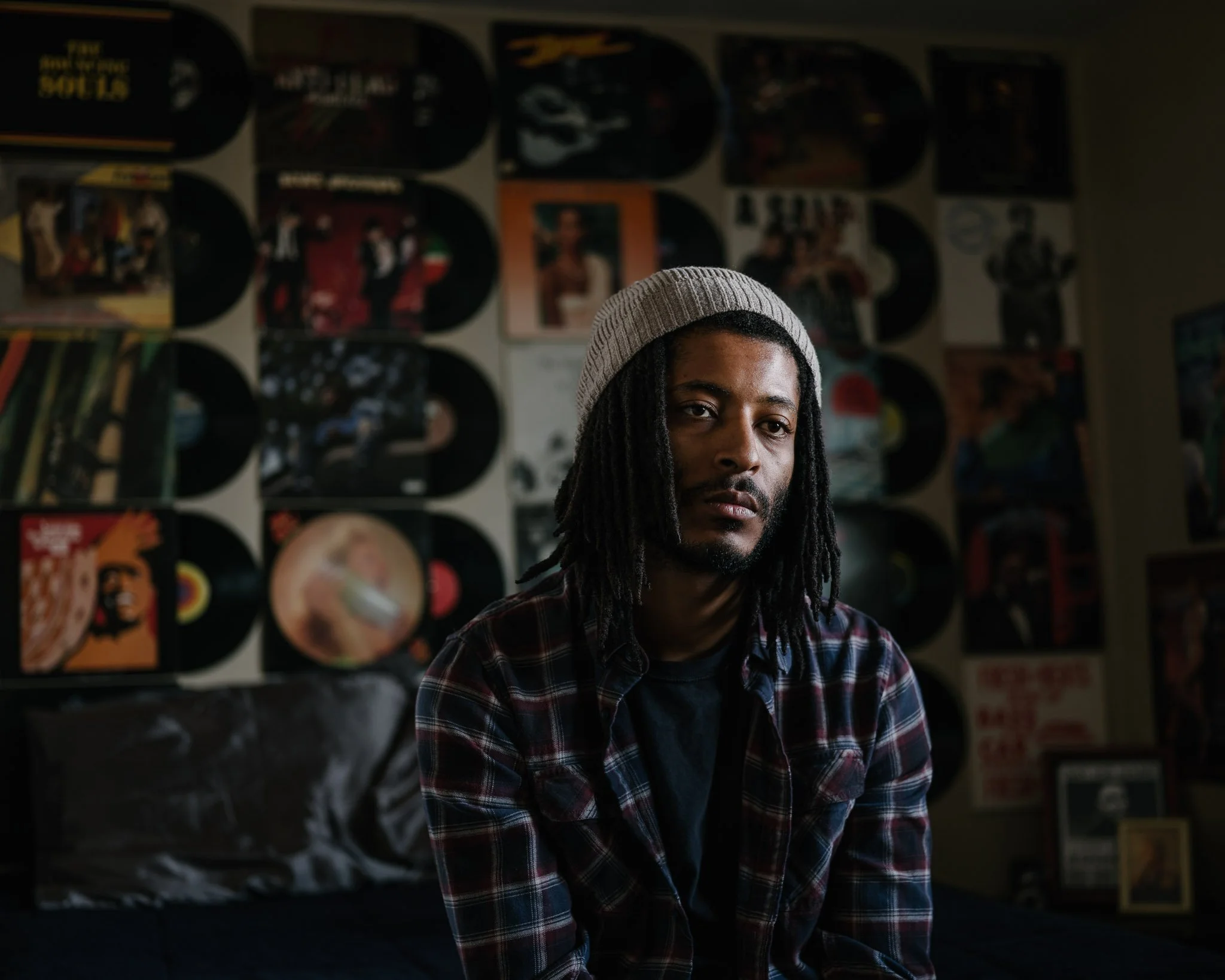 Handsome young black man with a short beard, dreadlocks and a beanie in a spot of light in his apartment, the wall behind him lined with vinyl records and album covers from the 70s and 80s. He speaks of dread and dark fatalism.