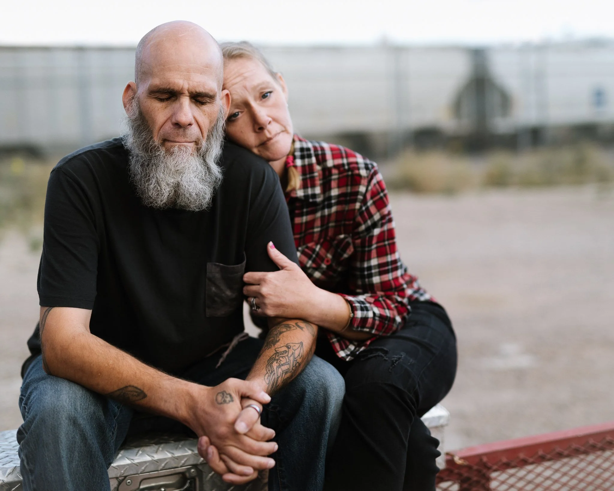 A bald homeless man and his wife sitting on a trailer in a train yard. His head is shaved and she has a long gray beard, his eyes closed. She sits behind him, her head on his shoulder, her hand on his arm.