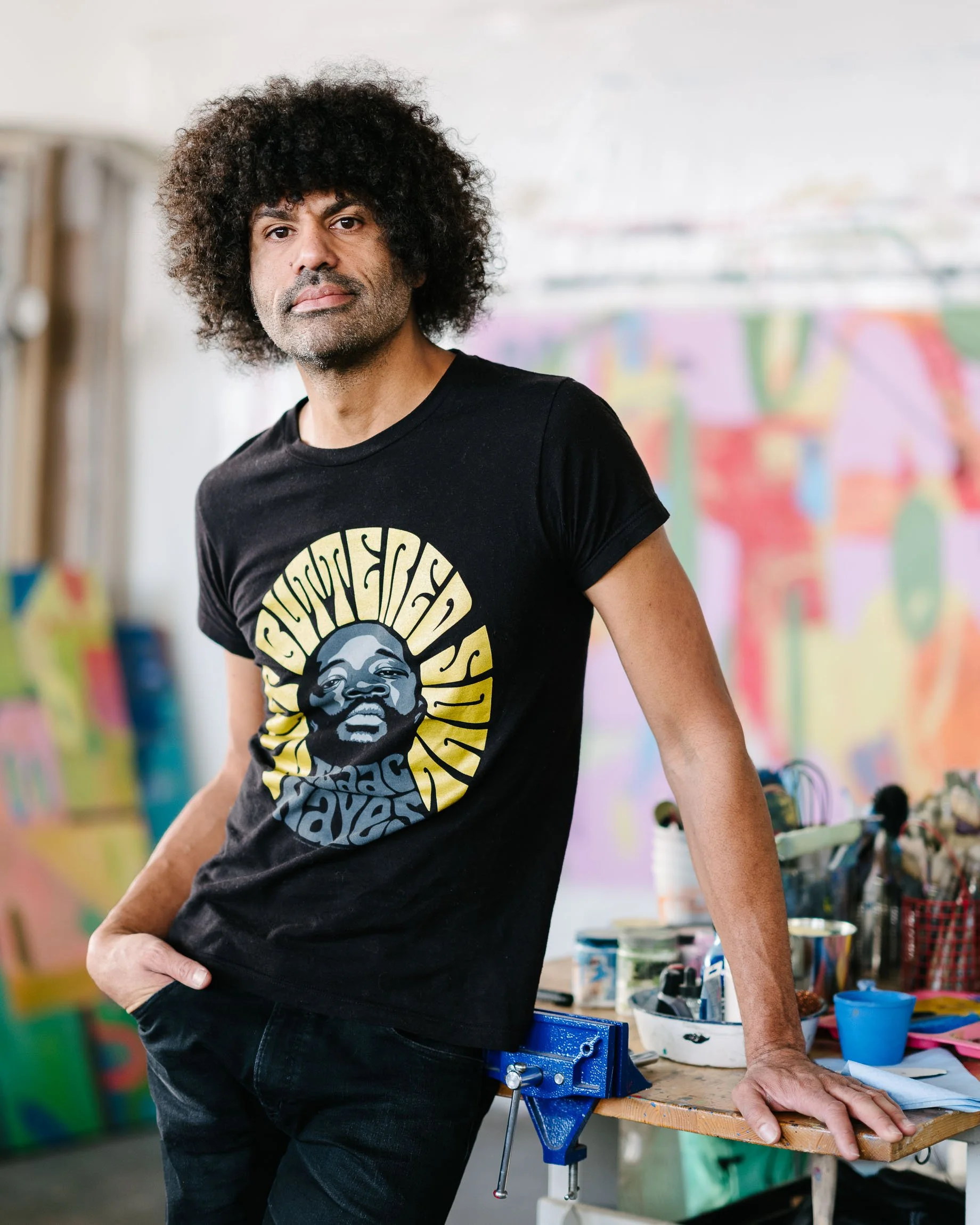 Denver artist MIchael Gadlin in his studio, languidly leaning against a table overflowing with painting materials, his work on the wall behind him.