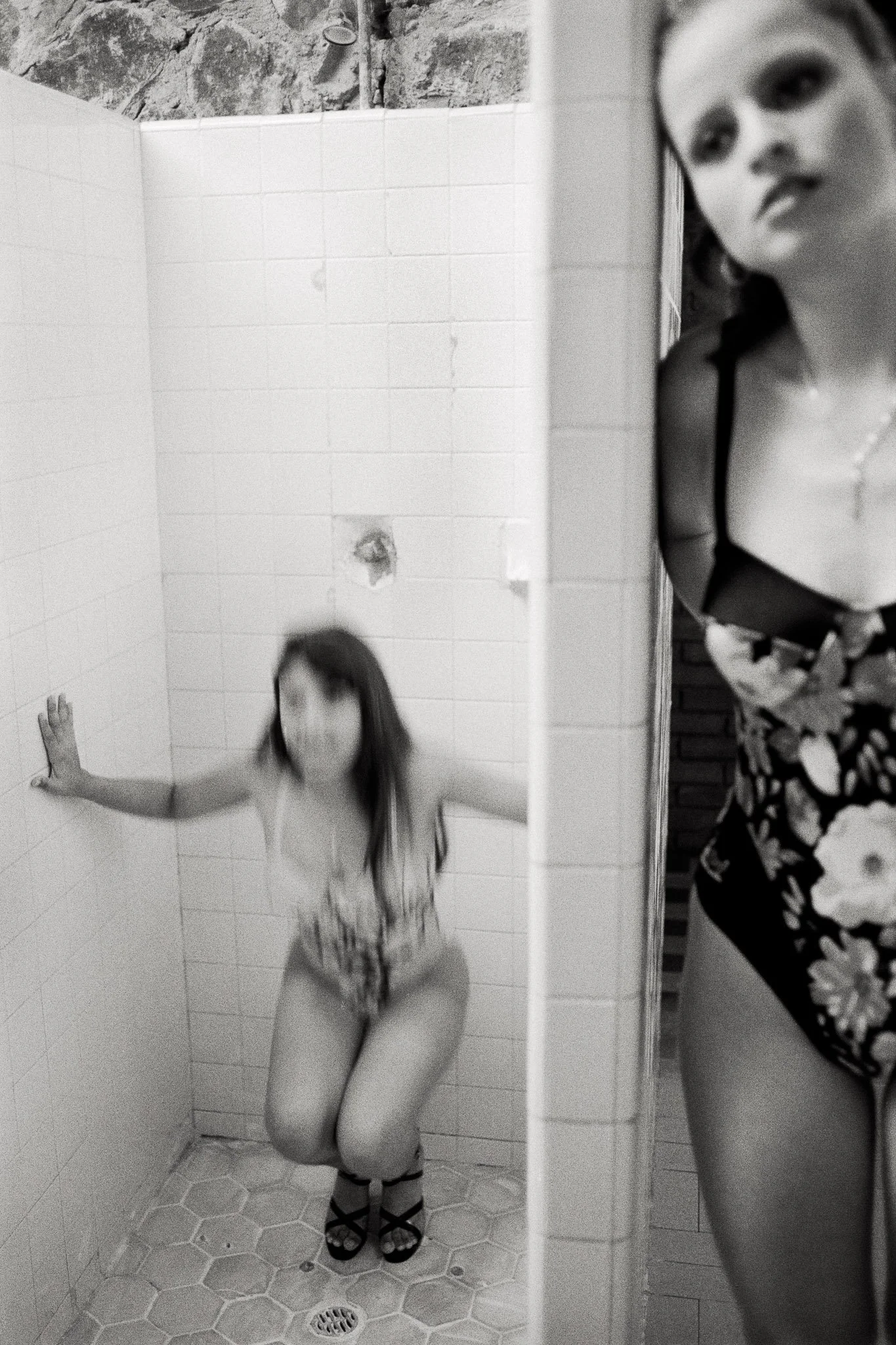In the shower stall of a swimming pool locker room, a beauty pageant contestant in a swimsuit and high heels does squats to firm her thighs and butt at the last minute before a public presentation.  A contestant in the foreground has a distant gaze.