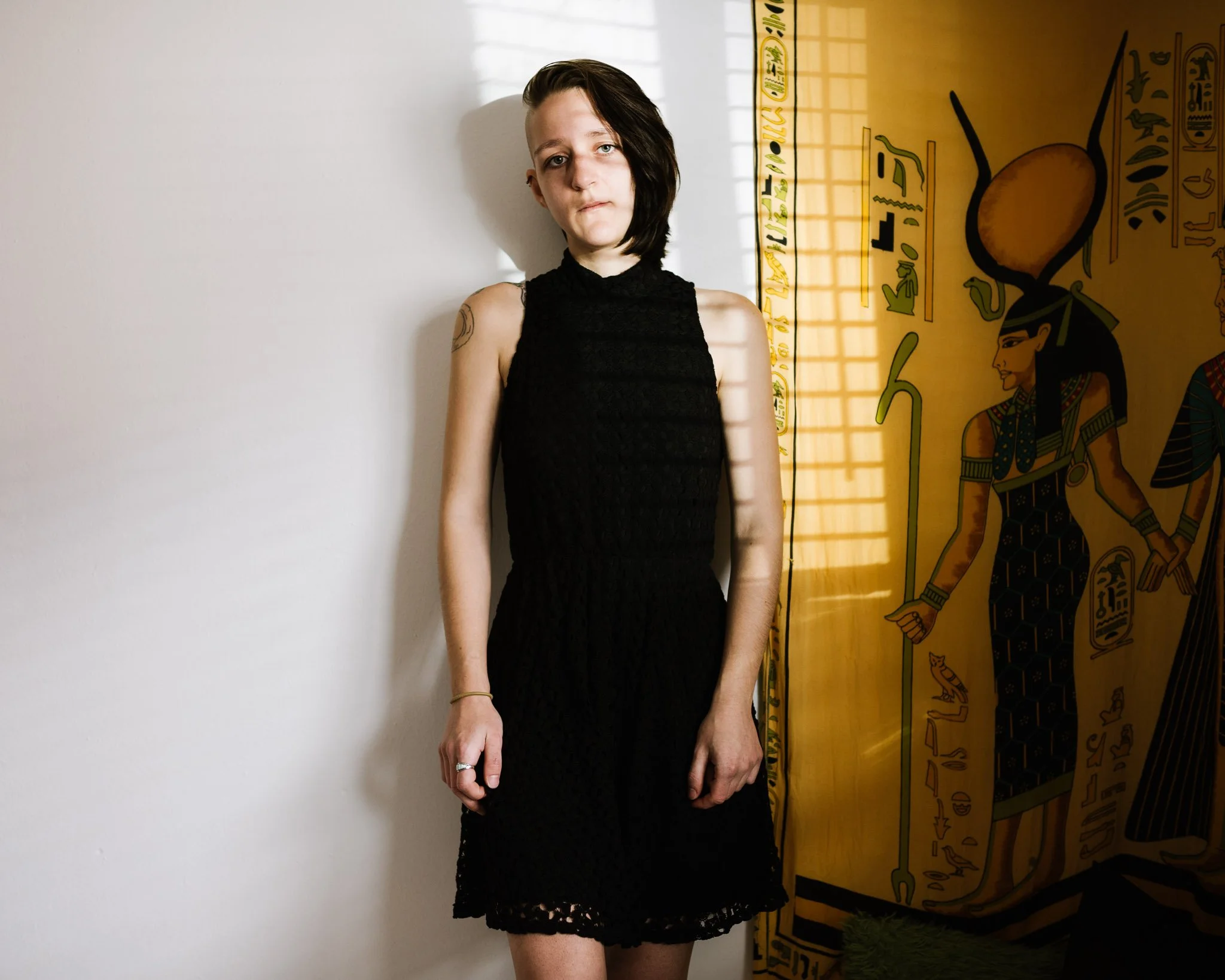 A pale white woman in her late 20s, her head shaved on one side, wears a sleeveless black lace dress as she stands in her room, sun shining through venetian blinds, a large yellow Egyptian tapestry on the wall.