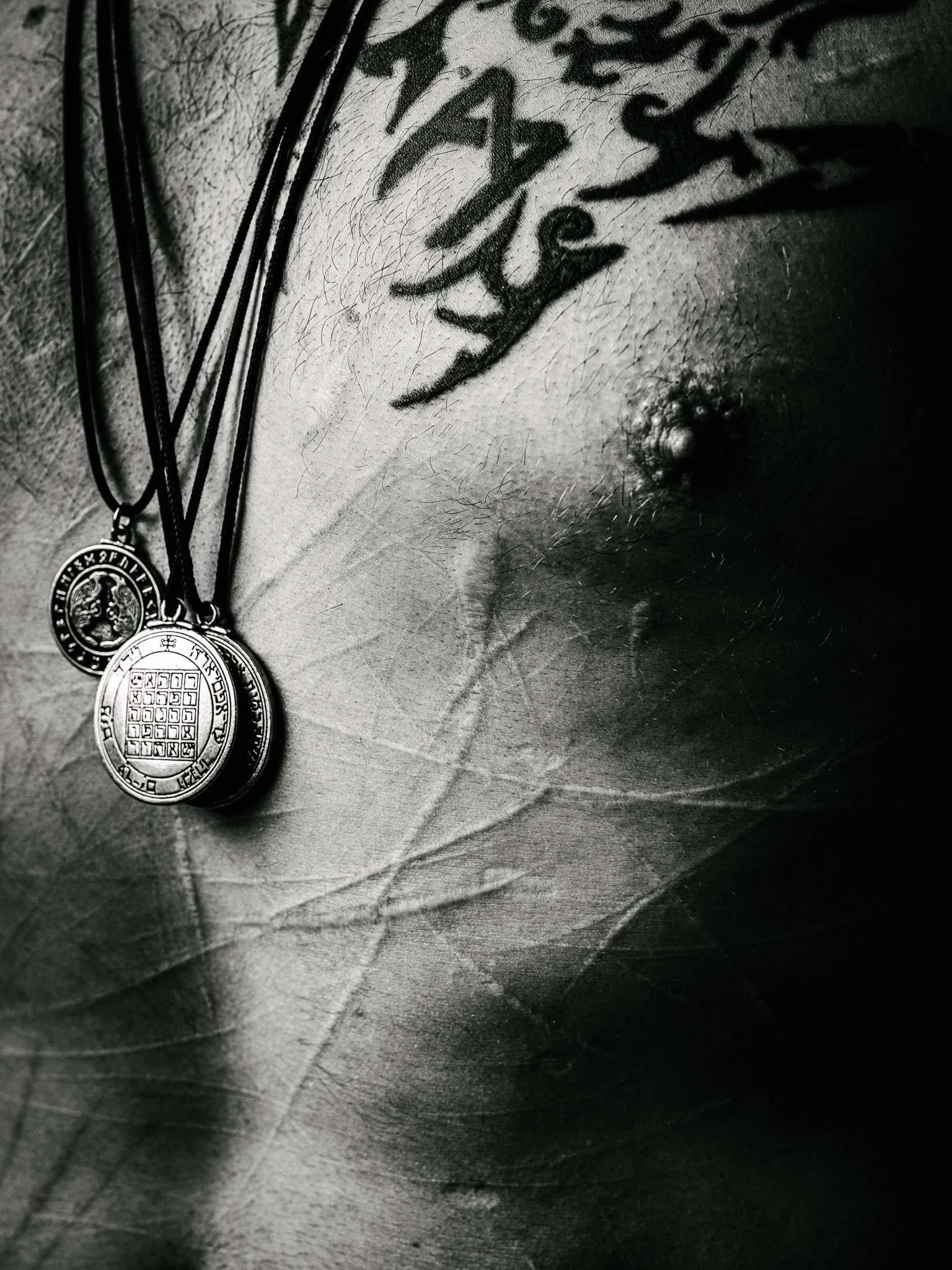 Black and white close up of the chest of a former Marine, raised welts and scars cover his chest from self scarification. Several pendants with runes on his chest, and black symbol tattoos.