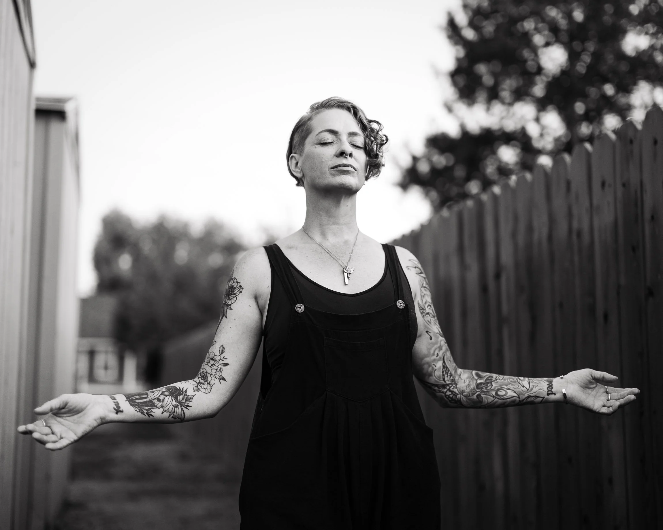 Monochrome image of a white woman with hear cropped on one side and a black jumper, outdoors between a shed and a wooden fence, her arms outstretched in prayer and meditation, tattoos on her arms, her eyes closed. Monochrome image with shallow focus.