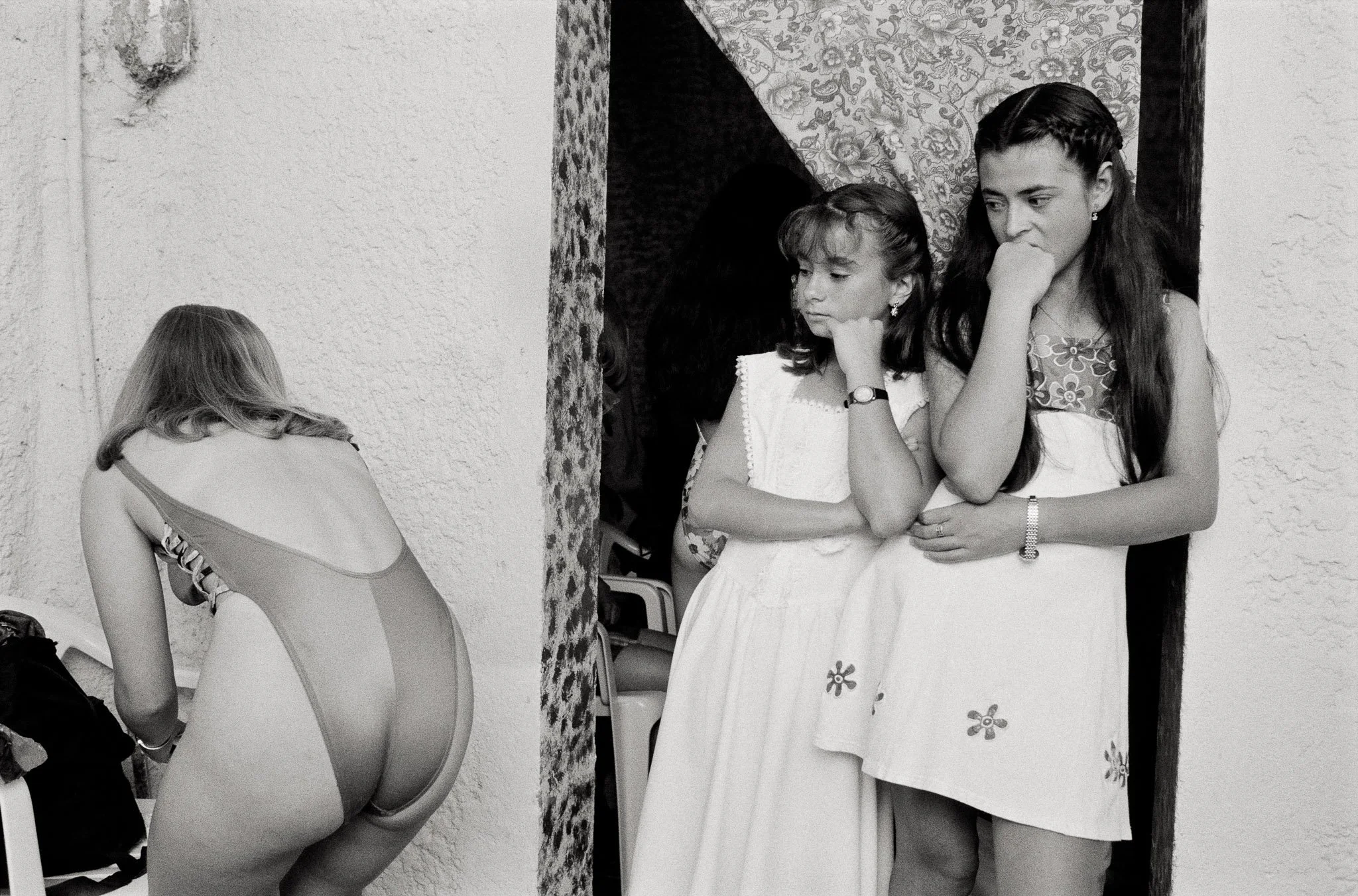 A beauty pageant candidate prepares in a changing room, her back to the camera. Two younger girls stand in an adjacent doorway, hands on chins, arms crossed in mirrored pose, looking at the queen's backside critically.