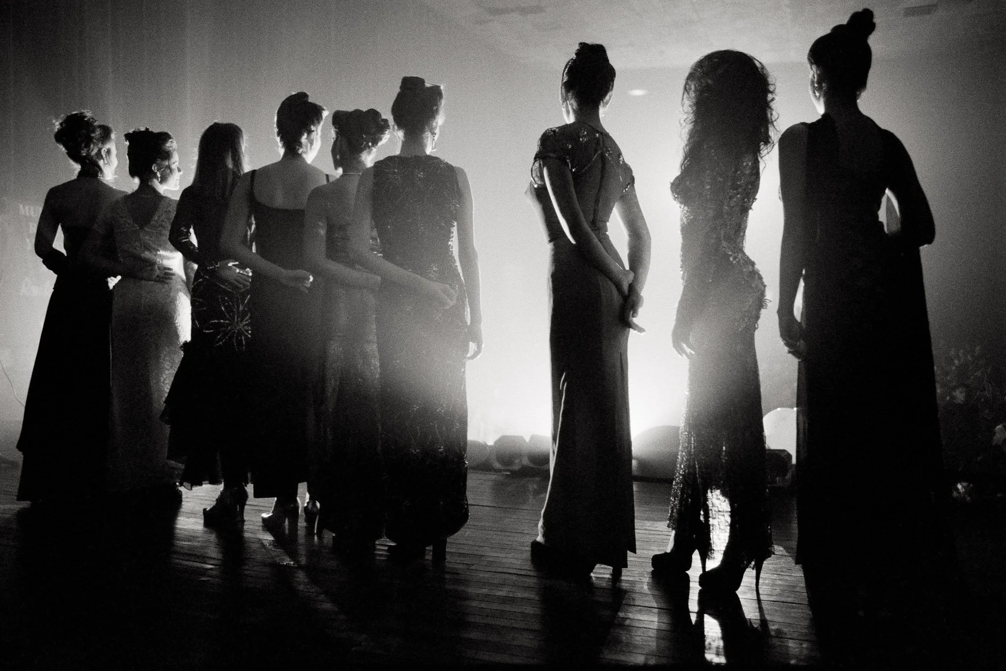 Black and white photograph of beauty queens lined up on a stage, backlit in a hazy auditorium.