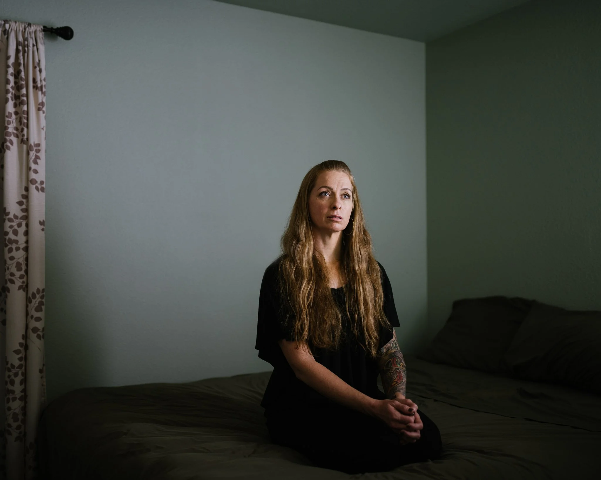Portrait of a woman kneeling on her bed in a dark room at night, hands clasped, long hair, in a small spot of light. She speaks of her fears and history of sexual violence.