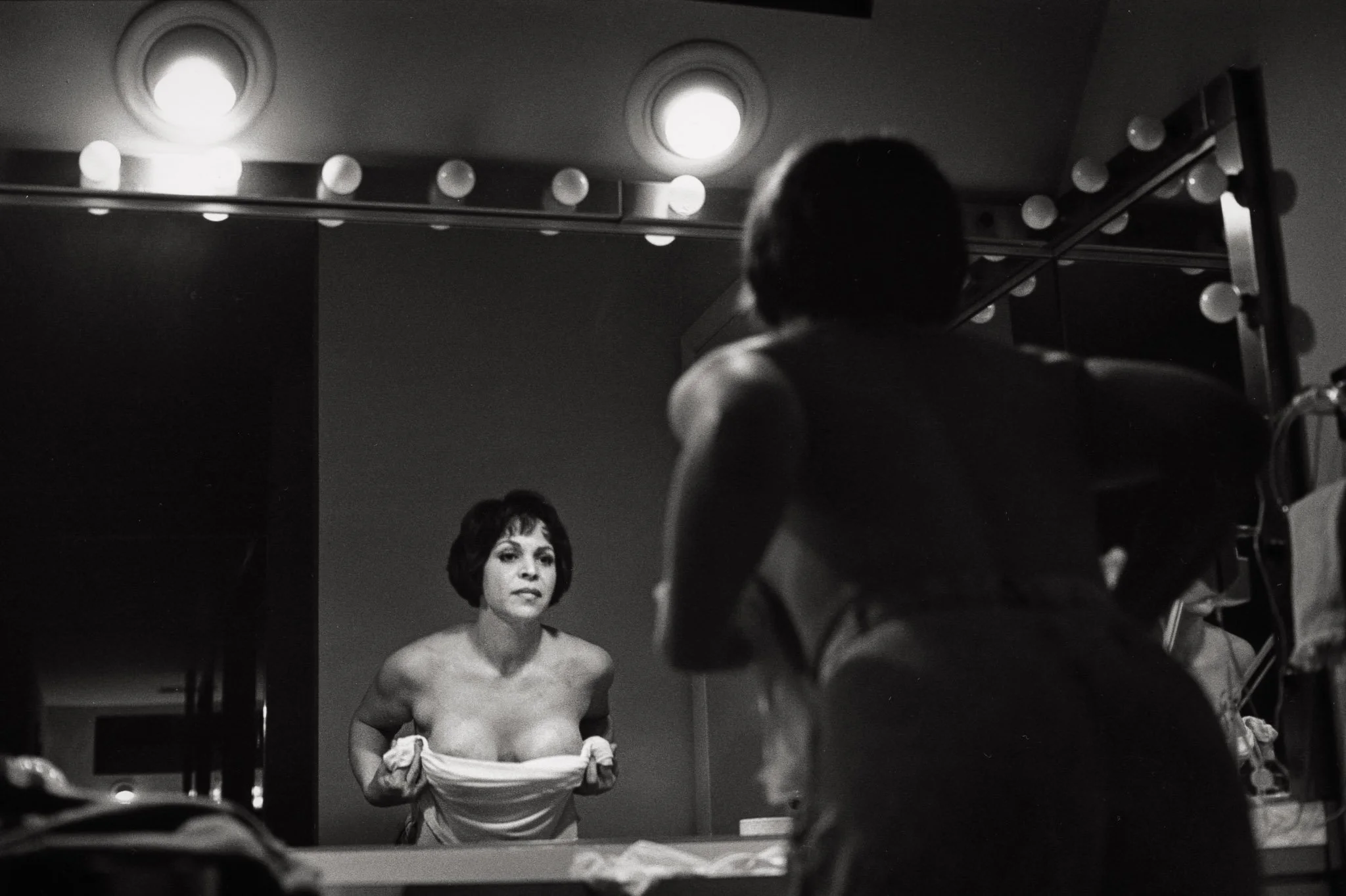 A woman leans forward in front of her bathroom mirror, a towel drawn across her chest, as she assesses her cleavage and nipples during her process of breast reconstruction following a mastectomy.