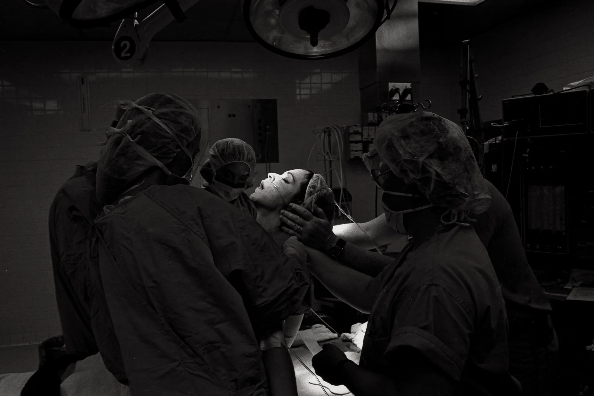 A woman's face is lifted into a tiny pool of light in an operating room, eyes closed, mouth open, while the room itself is very dark, the masked surgeons looking like passing ghosts around her, just barely visible.