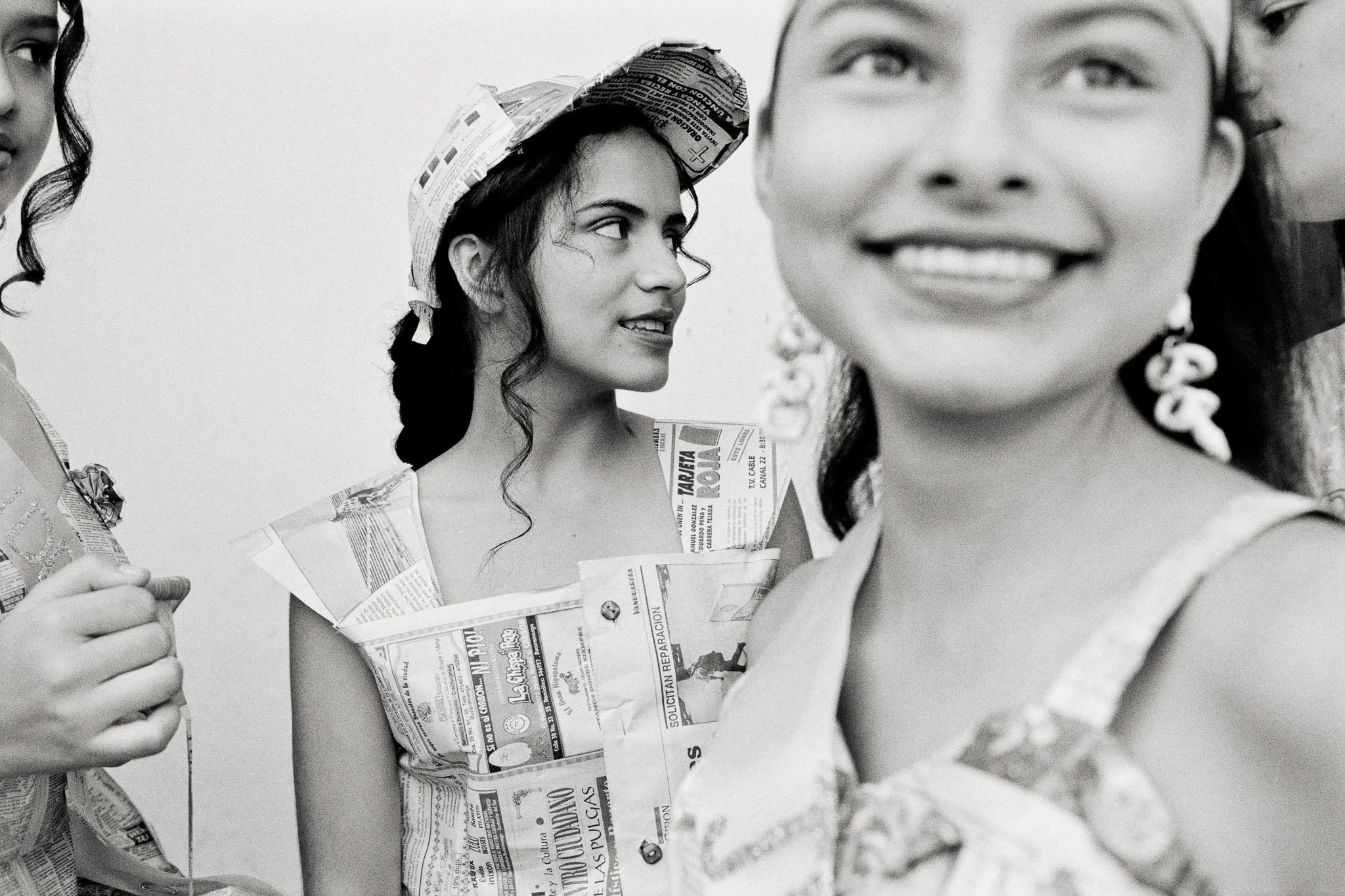 Graphic image of four young women dressed in outfits made of newspaper, tightly cropped, some faces on the far edge of the frame, four distinct expressions in a tight composition, high-key.