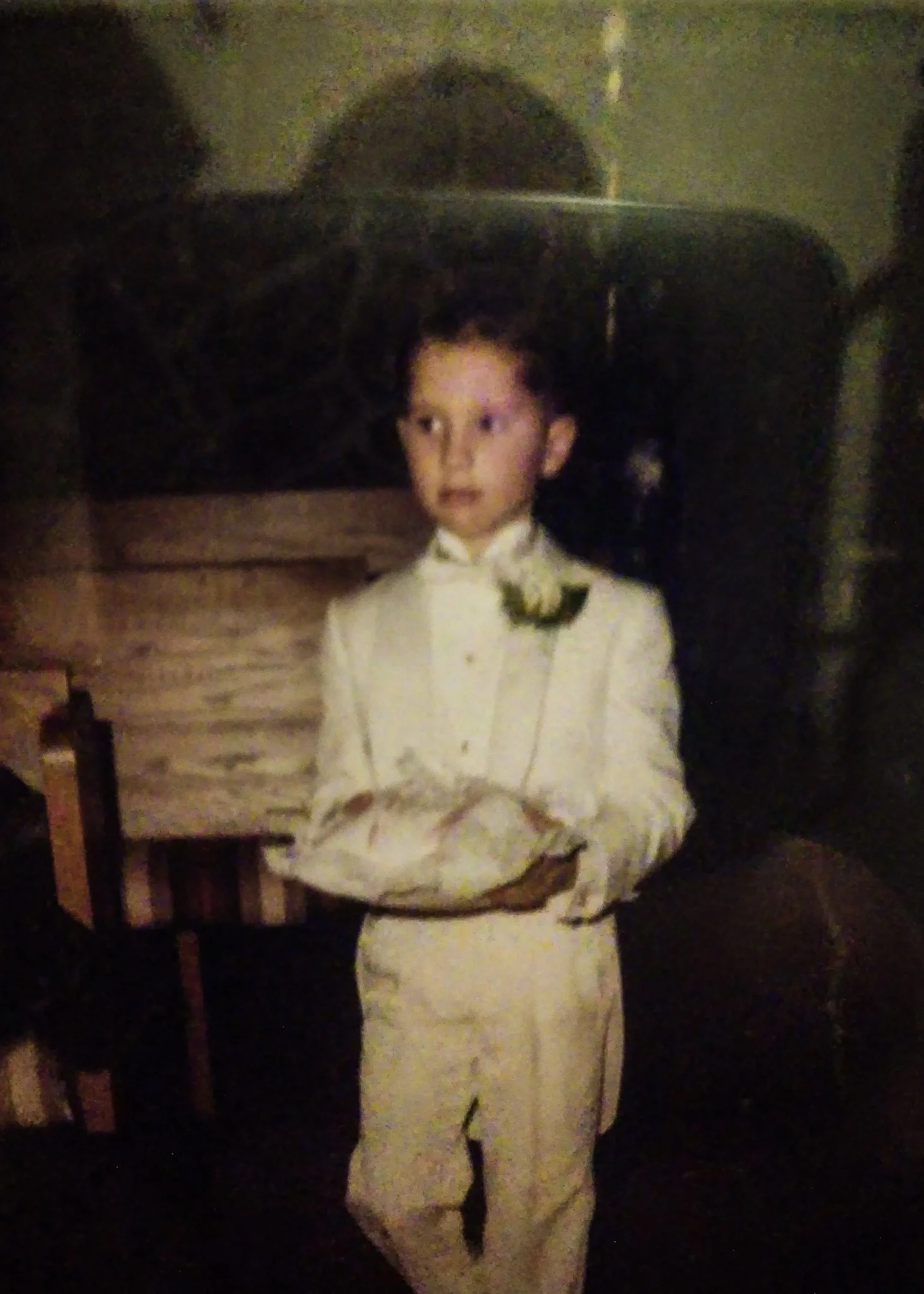 Vintage photograph of a young boy serving as a ring bearer. He is dressed entirely in white, with a corsage. Grainy, blurry photo taken with direct flash.