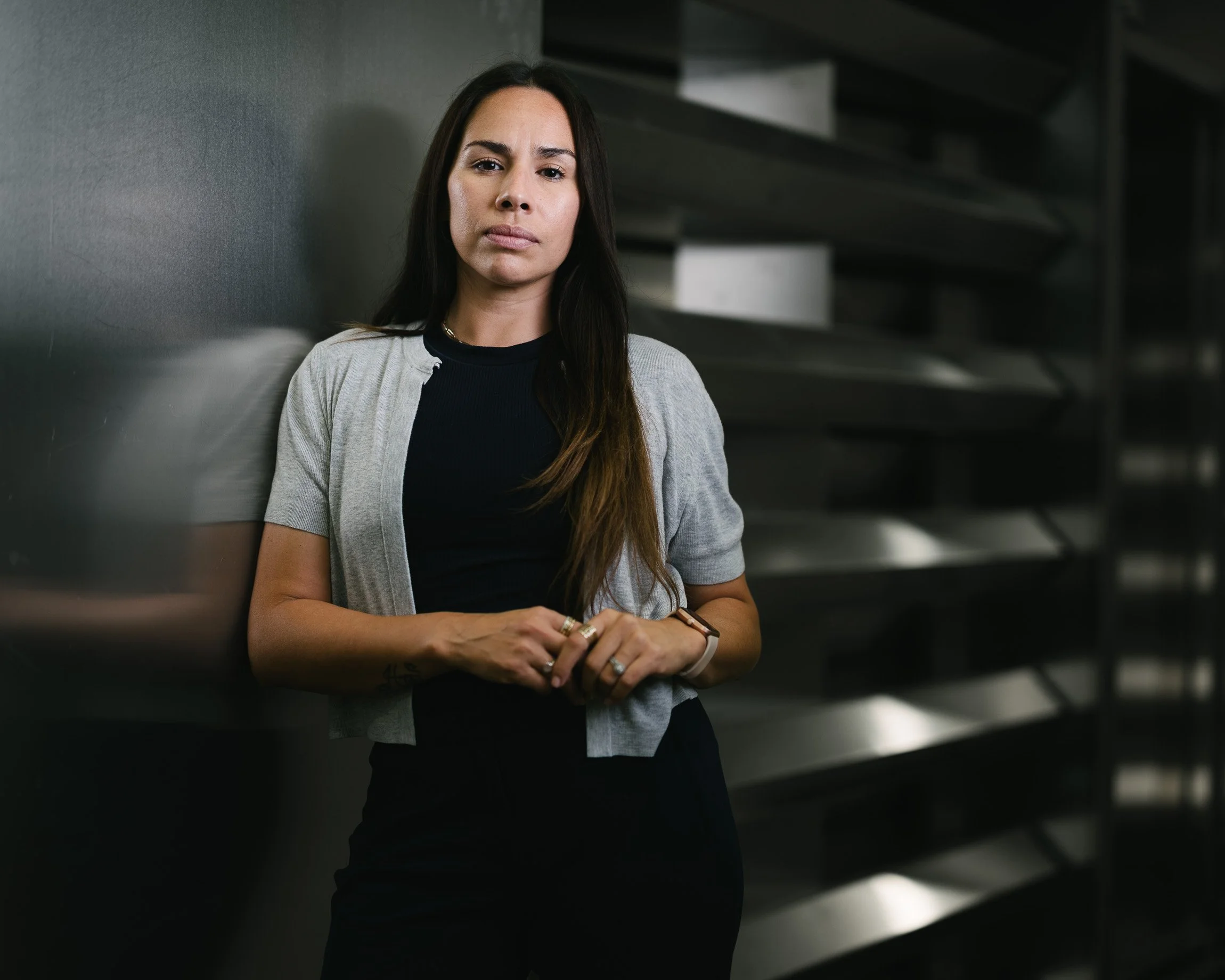 Latina woman in black with a gray sweater looking at camera with strength and resolve. The background is dim, metallic and industrial.