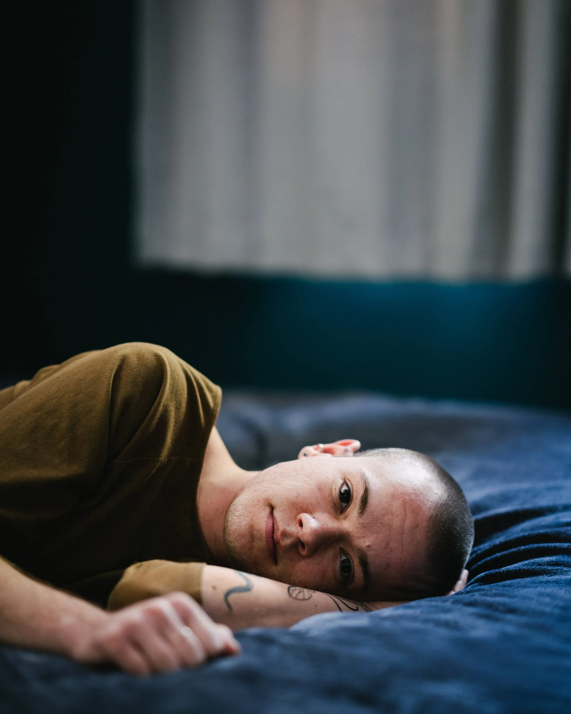 A young man in his 20s with shaved head and a gentle demeanor lying on his blue bedspread in a room with blue walls and white curtains, cradling his head in his hand, his other hand on the bed, in a gentle pose.