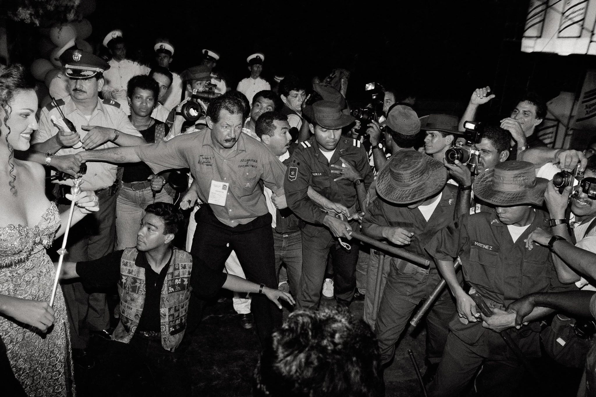 A newly crowned beauty queen crammed into the far edge of the frame faces a chaotic scrum of photographers and onlookers held back by military police with batons. Security officials and officers with walkie talkies try to manage the chaos.