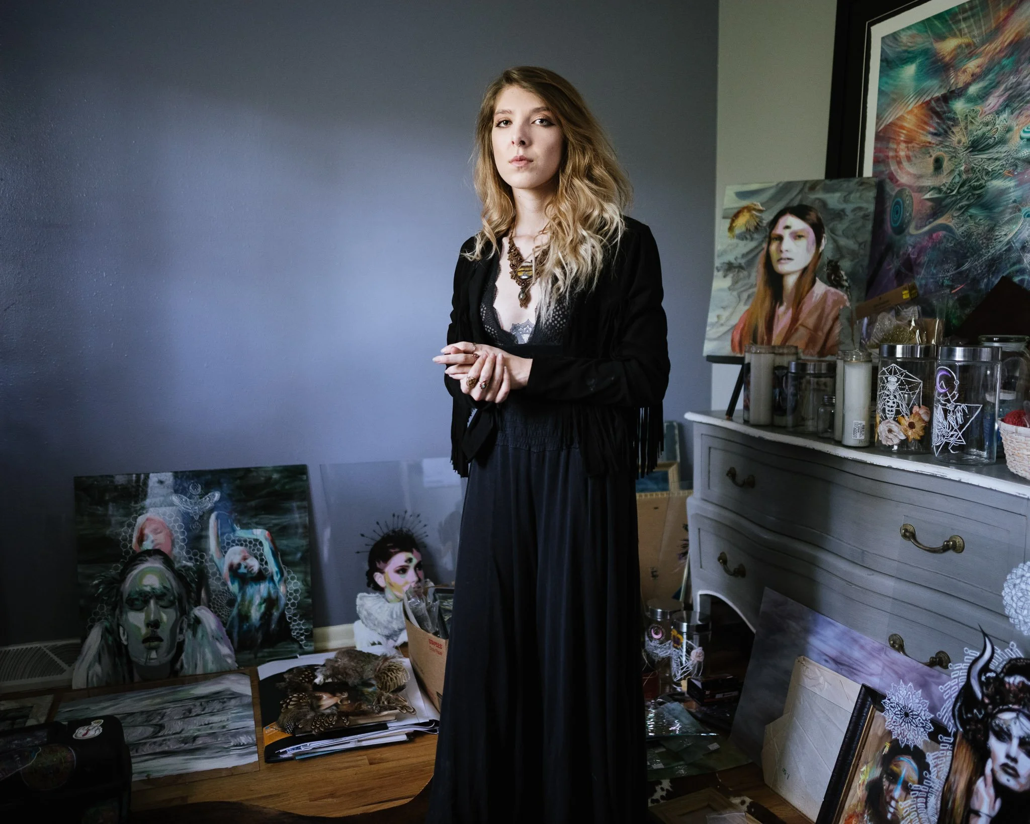 Blue-toned image of a woman in a long black dress with a low neckline, tattoos and jewelry, with a witch-like persona. She is surrounded by her glass paintings prompted by recurring haunting dreams.