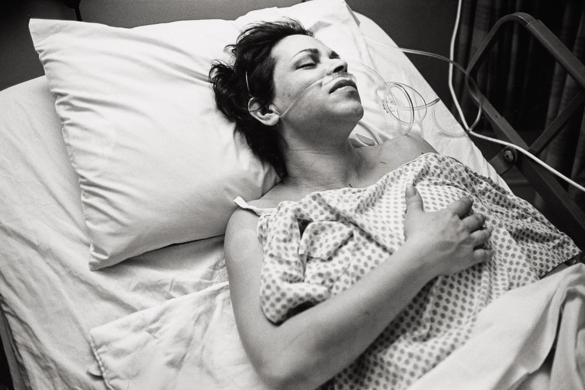A woman lies in a hospital bed following a mastectomy, her hand feeling the place where her breast used to be, relieved by its absence.
